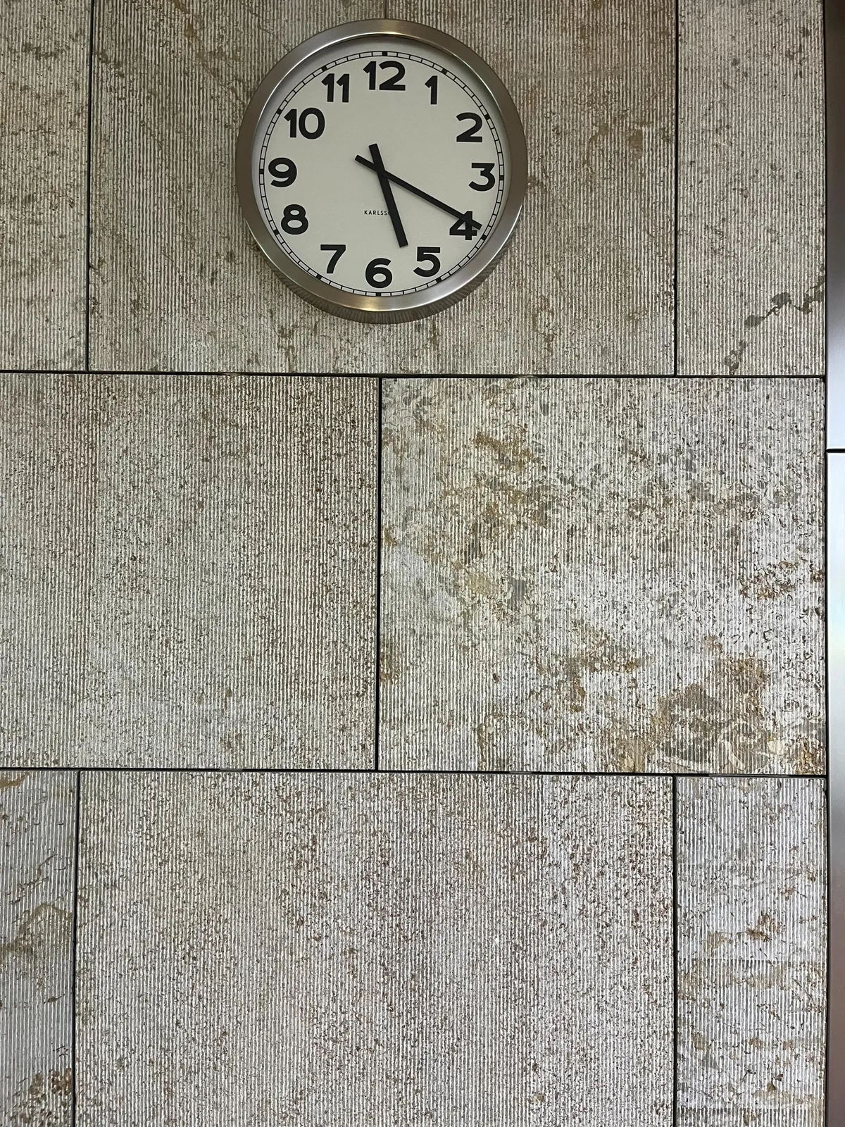 A modern clock on a modern, gray, stone wall. It points at 19 minutes past 5.