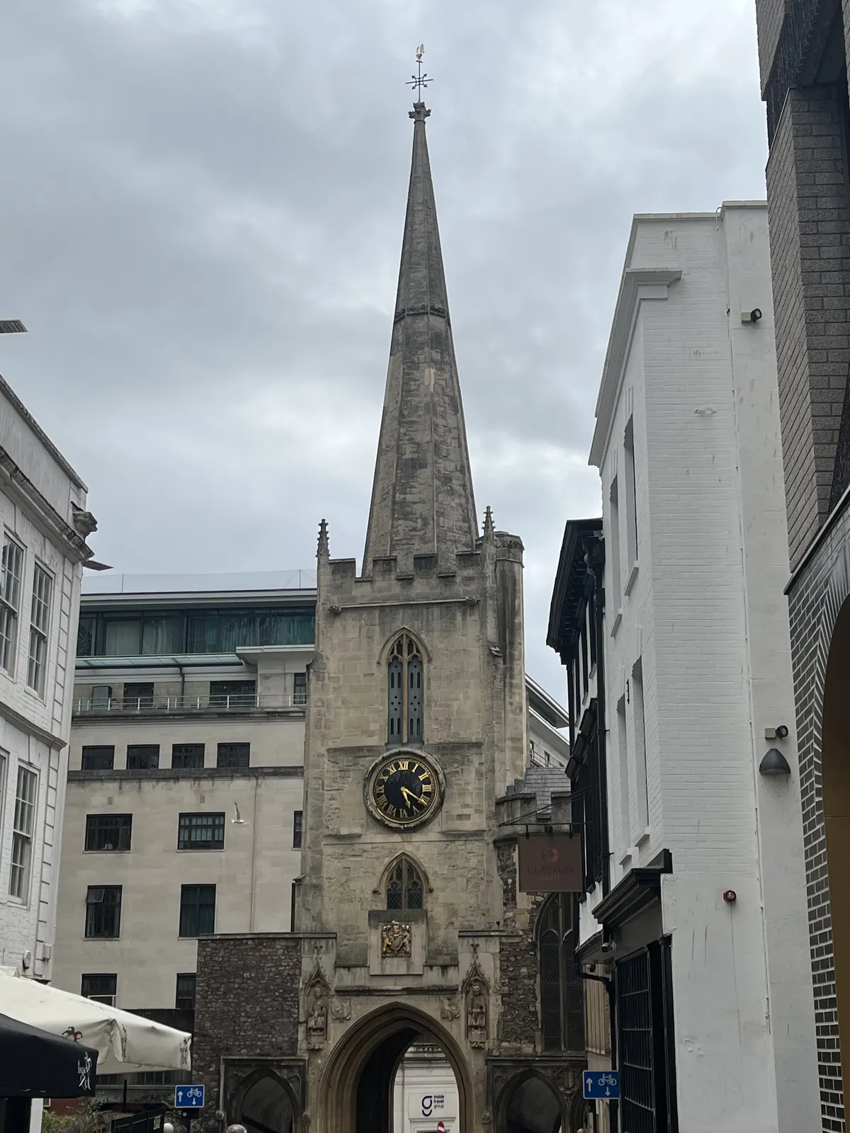 An old, churchlike tower with a gate in it, in an urban environment, surrounded by both old and newer buildings. The clock on the tower points at 20 past five.