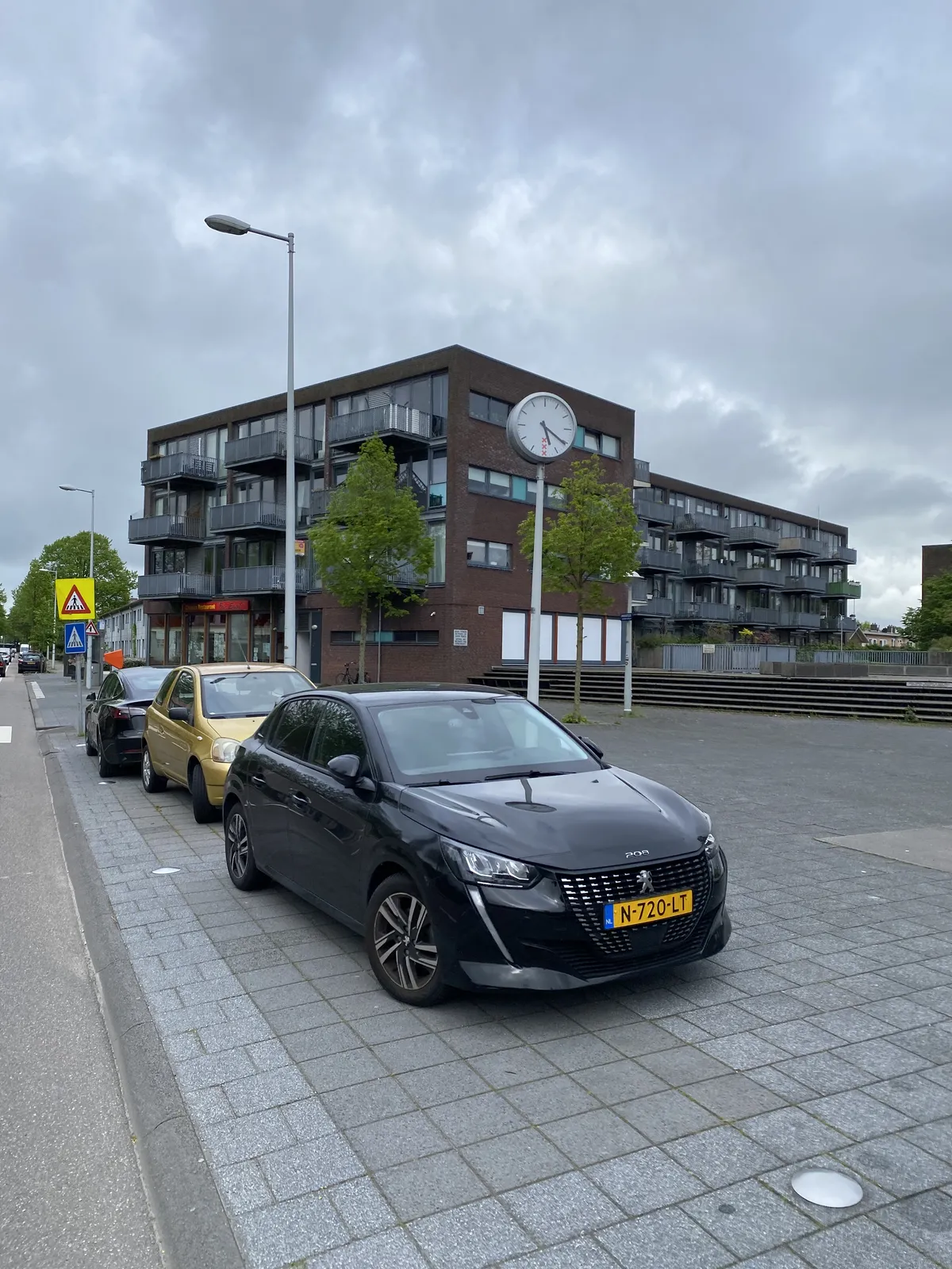 Cars are parked in front of a flat. There’s a clock on a pole that points at 21 minutes past 5.