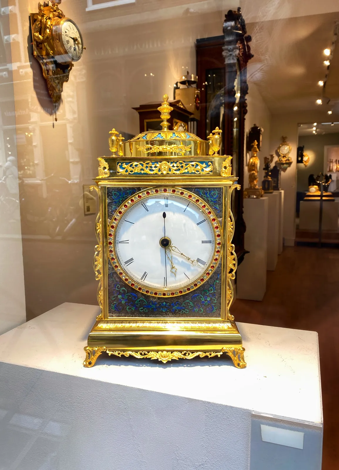 A richly decorated gold and blue clock, with some colourful gems. It’s displayed in the window of a clock shop. It points at 22 minutes past 5.