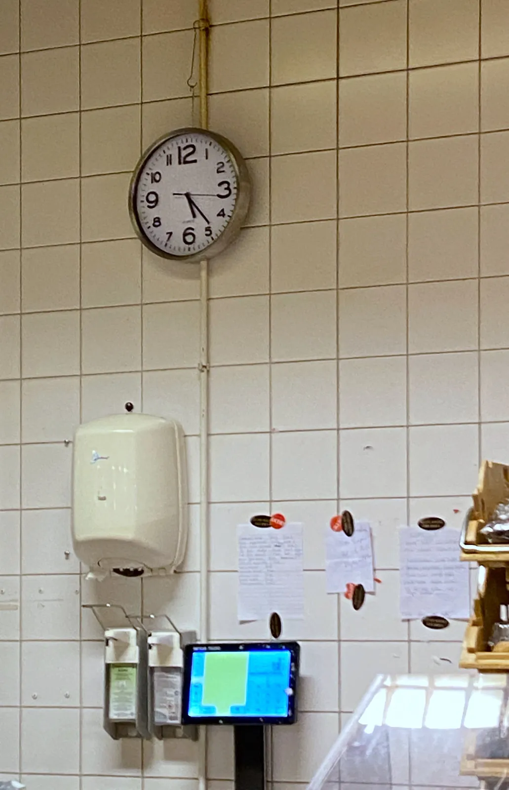 A wall of white tiles with a dispenser for paper towels, two soap dispensers, an interactive display and a clock. The clock points at 23 minutes past 5.