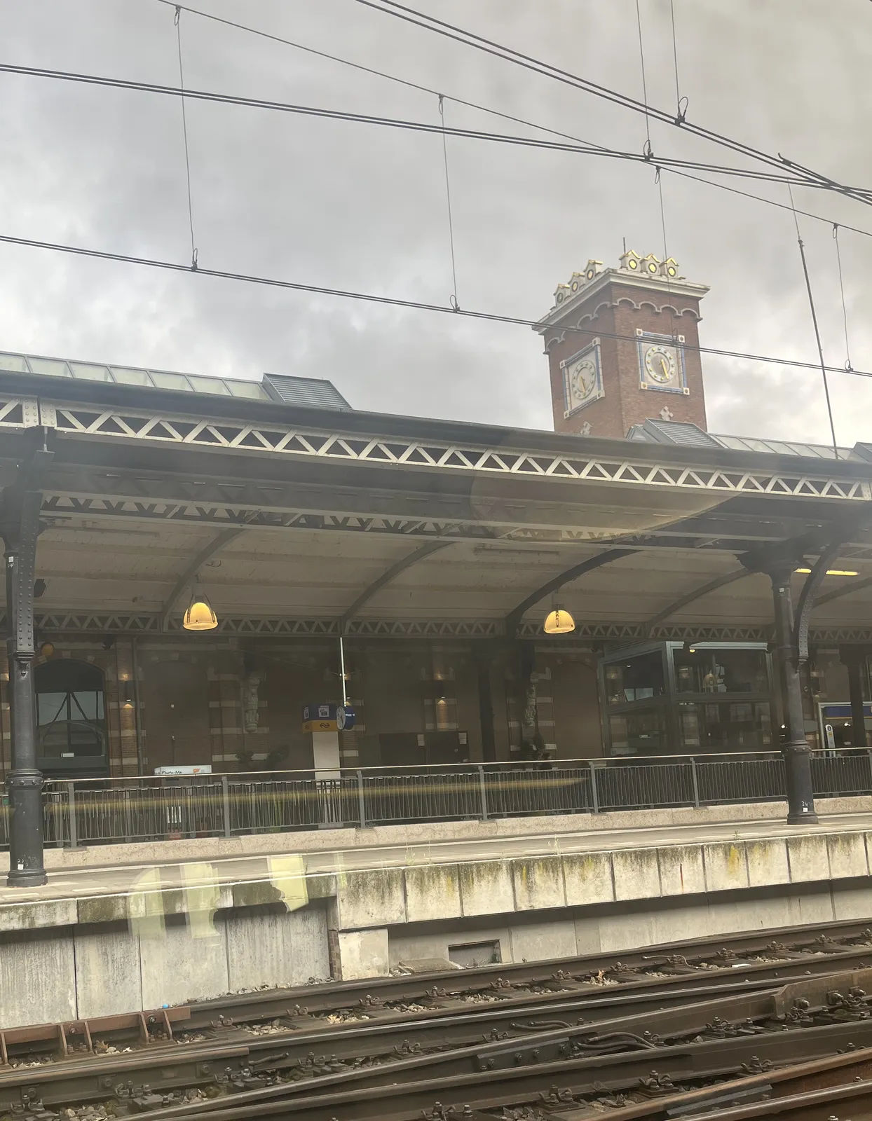 A picture taken at a train station. There are train tracks in the foreground, a platform behind it, and above the roof of the platform towers a monumental, yet colourful tower with a clock on both visible sides. Because of the distortion caused by perspective they both seem to point at slightly different times. The one on the left seems to point at 29 minutes past five, and the one on the right at 25 past five.