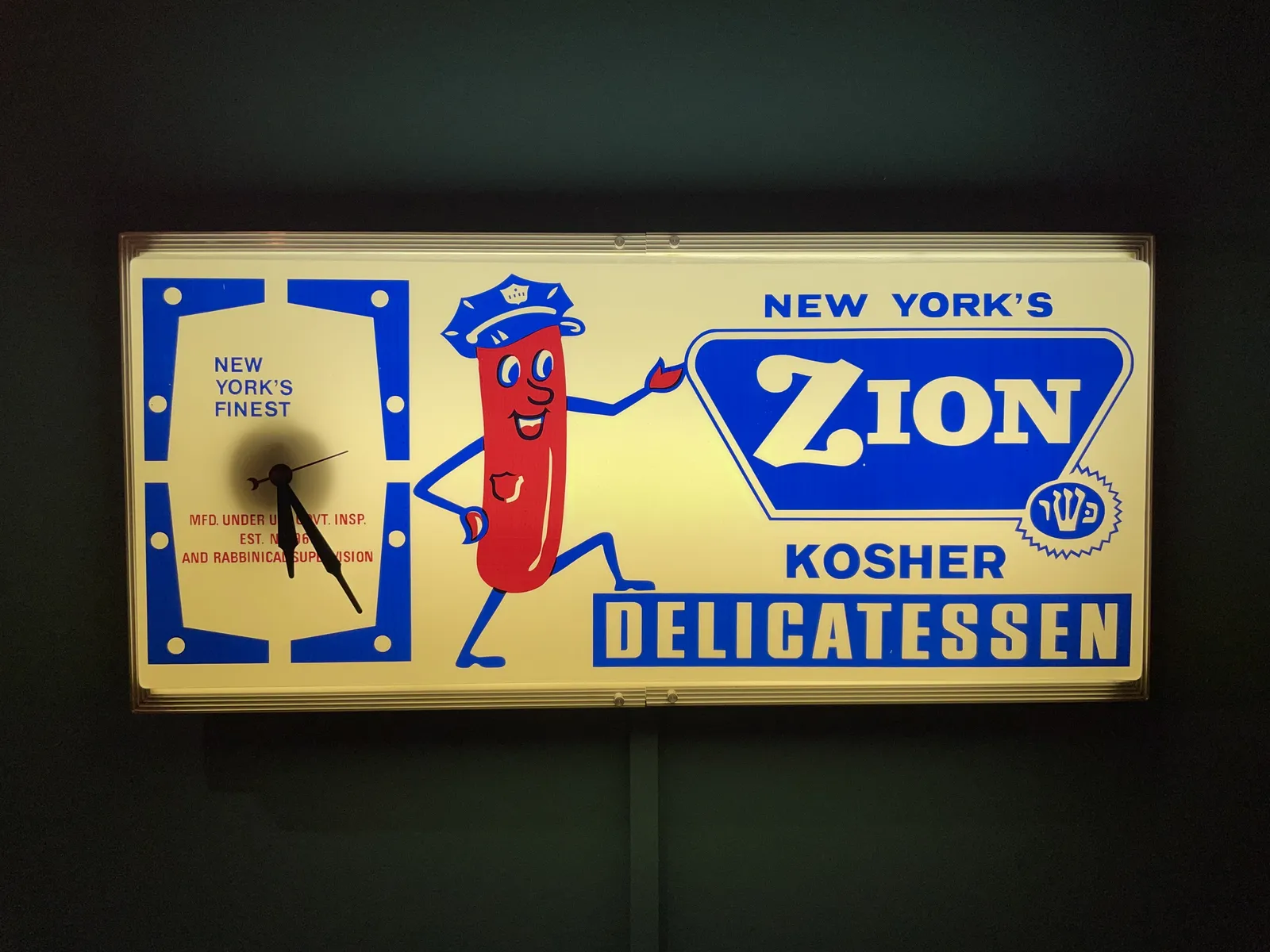 A lit sign, with a happy sausage wearing a police cap. It’s a sign for New York’s Zion kosher delicatessen.  On the left of this sign there’s a clock, that says it’s New York’s finest, with a message about rabbinical supervision. This clock points at 25 minutes past 5.