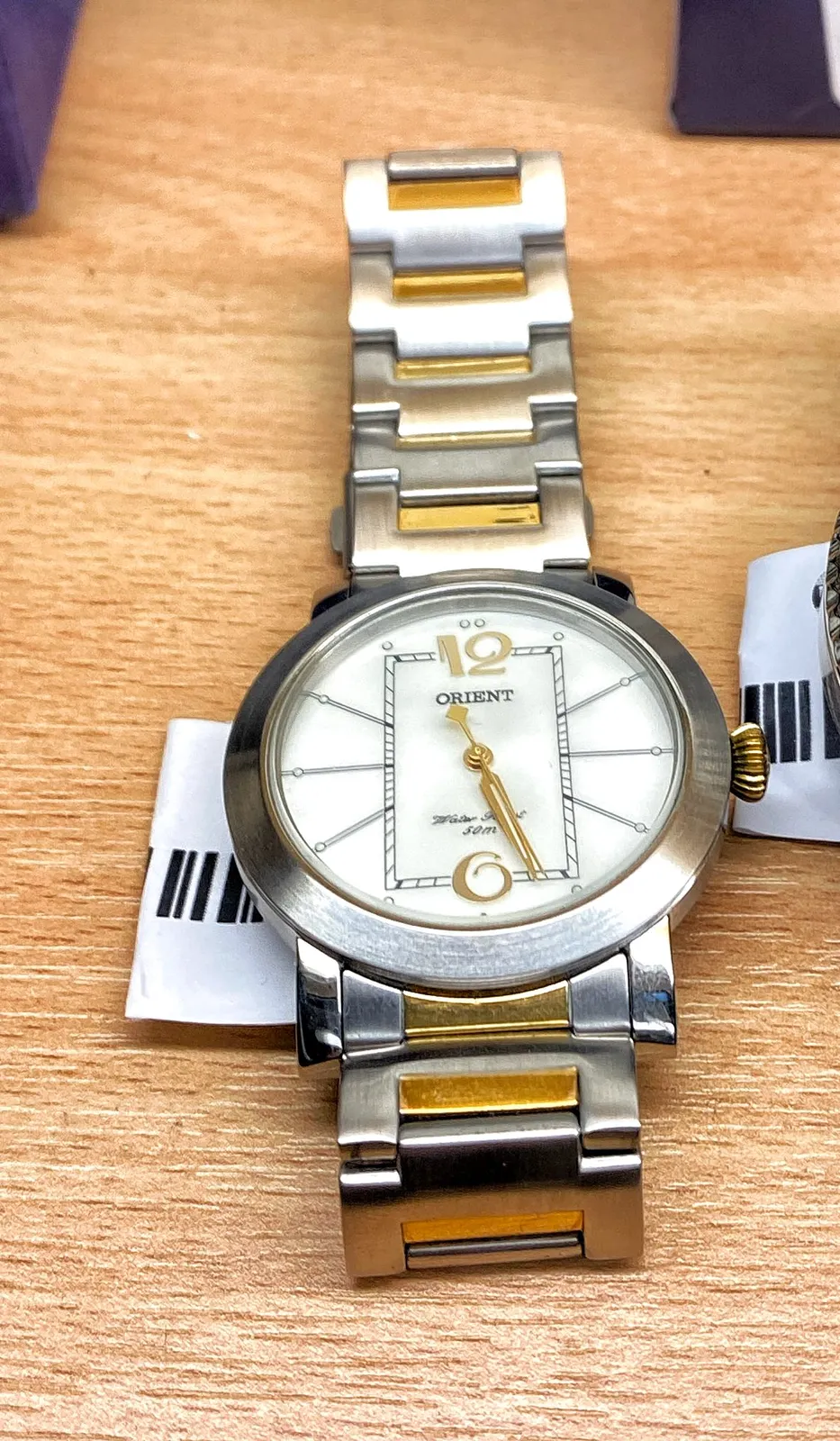 A silver coloured metal watch. It says Orient and something that’s covered by one of the hands. It points at 26 minutes past 5.