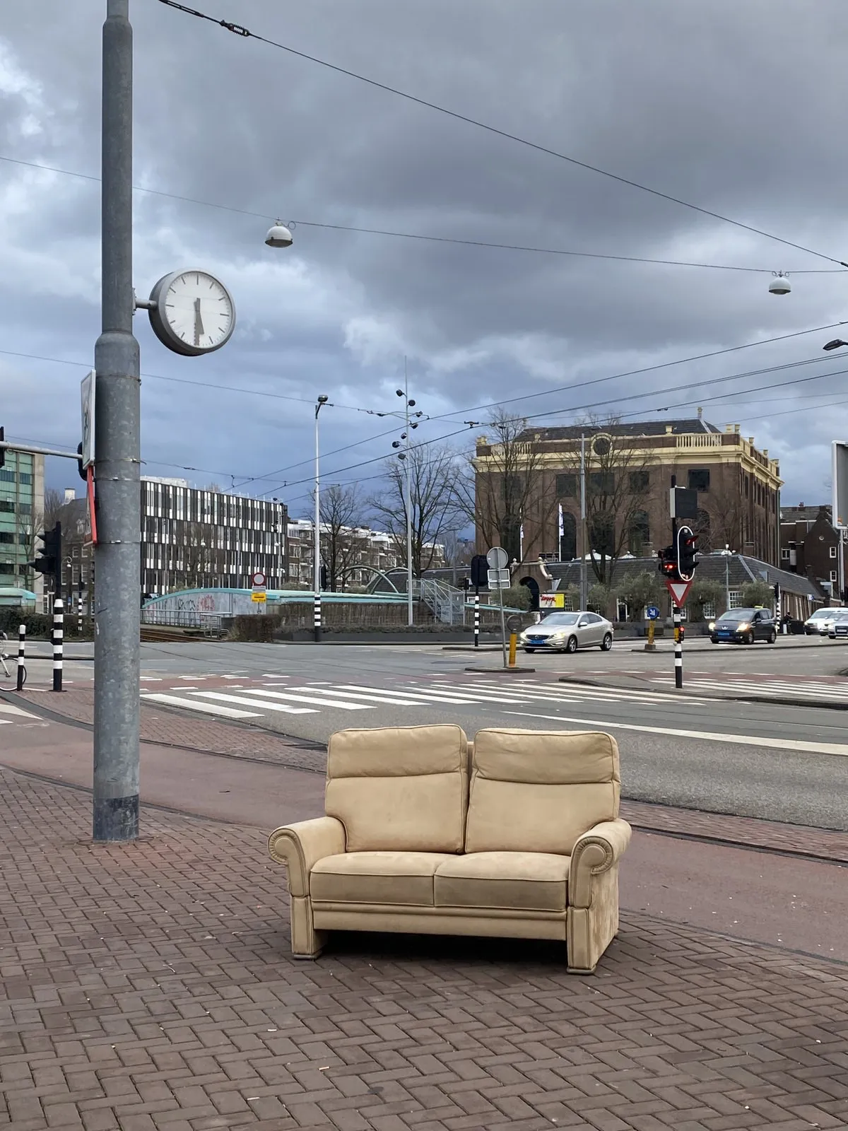 A picture taken on the Meester Visserplein in Amsterdam. In the background you can see the old Portuguese Synagogue. There’s a comfy looking couch in the foreground. Next to it is a pole with a clock attached to it which points at half past six.