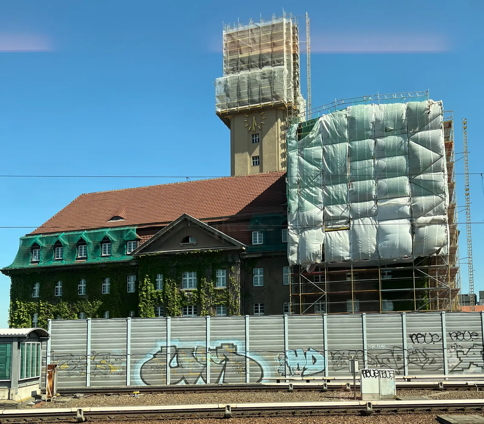 A building, in part in scaffolding. It has a tower with a clock on it, even thouhg it doesn’t necessarily look like a church. It points at 29 minutes to 6.
