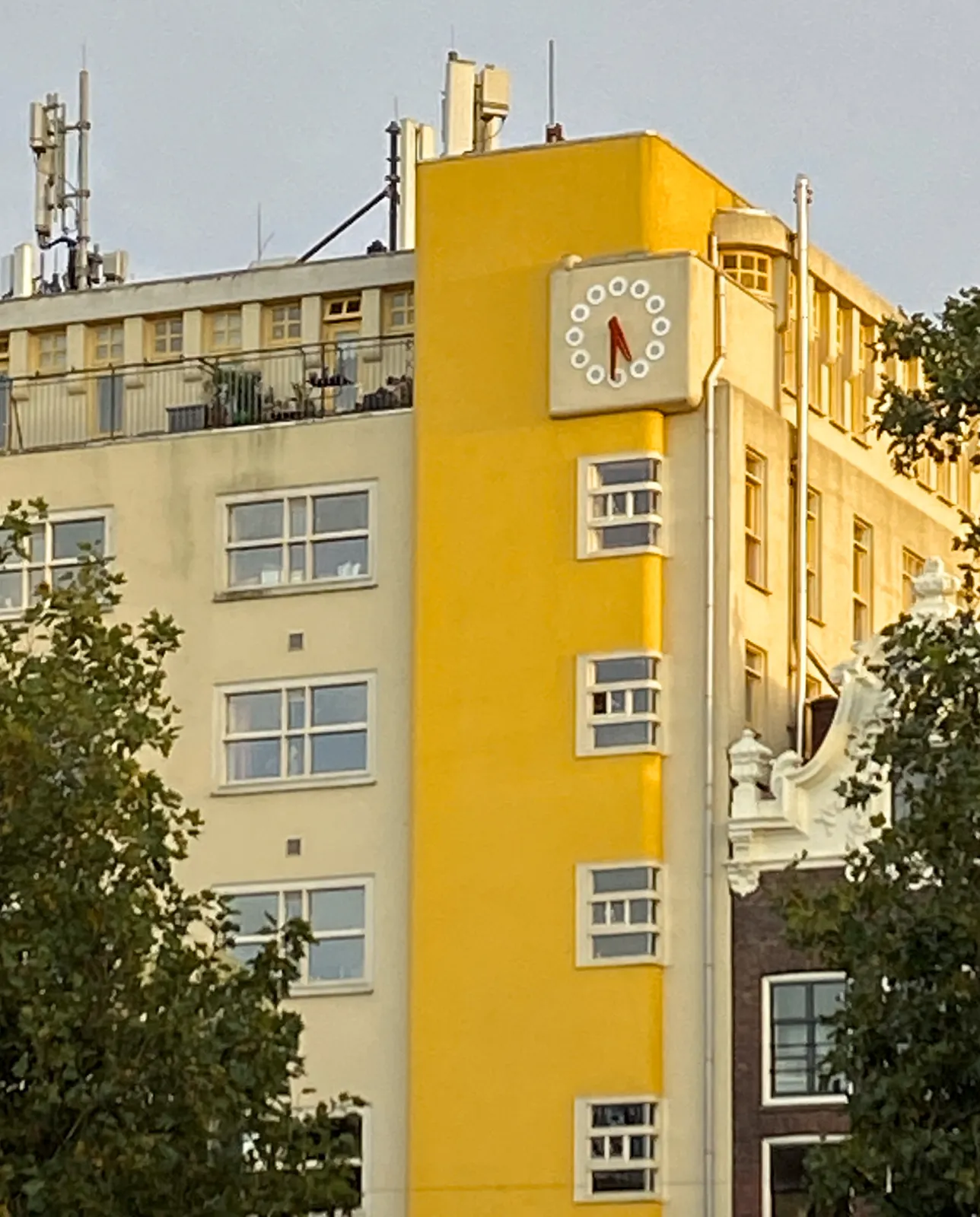 A striking, cream coloured modernist building, with a bright yellow column on its corner. At the top of this column there’s a clock with red hands that points at 31 minutes past 5.