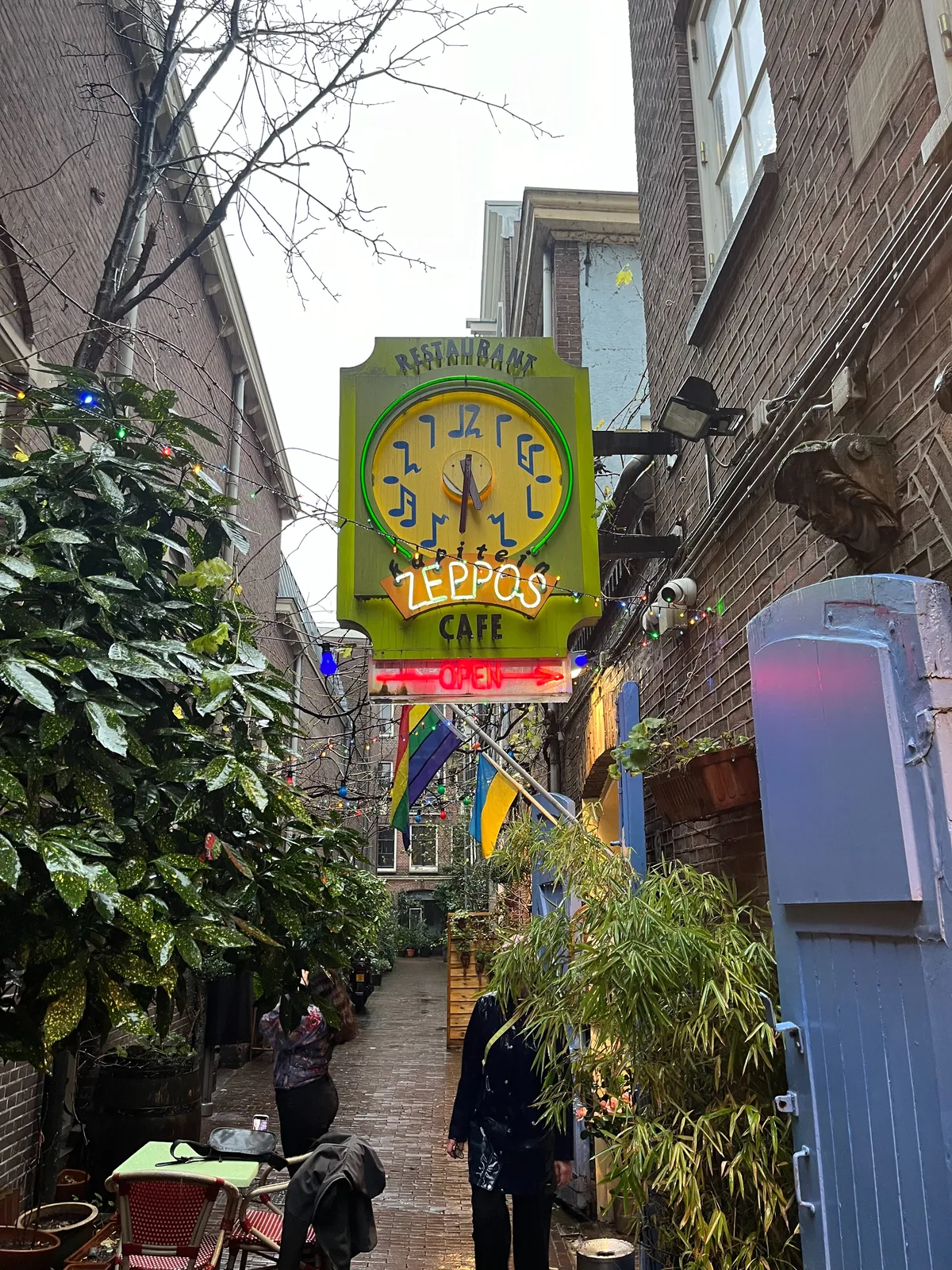 In an alley there are plants, rainbow flags, and a very colourful sign for restaurant kapitein zeppos. A red neon sign indicates that it is open. On the colourful sign there’s also a clock that points at 28 minutes to 6.