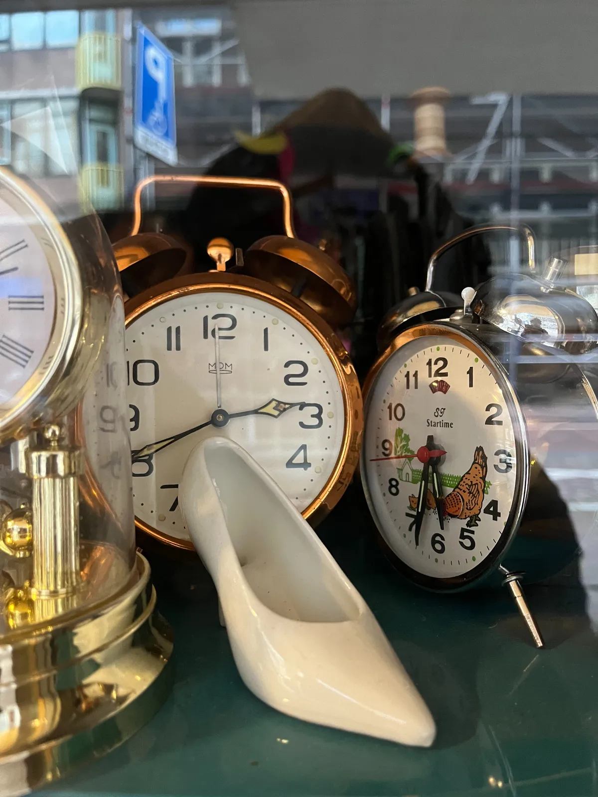 There’s a white porcelain shoe in the foreground. Behind it there are two clocks. The one on the right points at 33 minutes past 5. The one on the left points at the wrong time.  