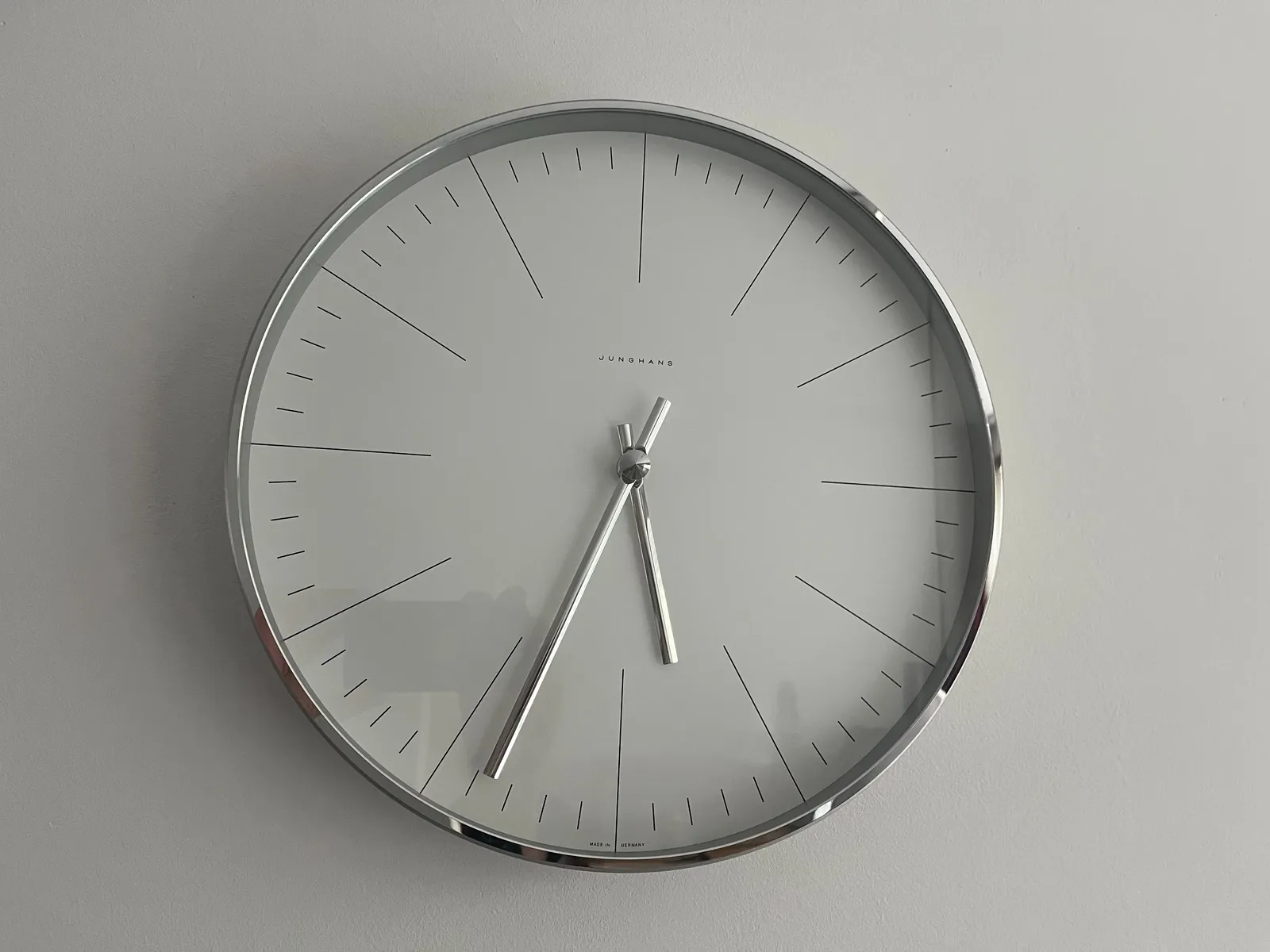 On a white wall is this rather minimalist white clock with silver hands. It points at 33 minutes past 5.