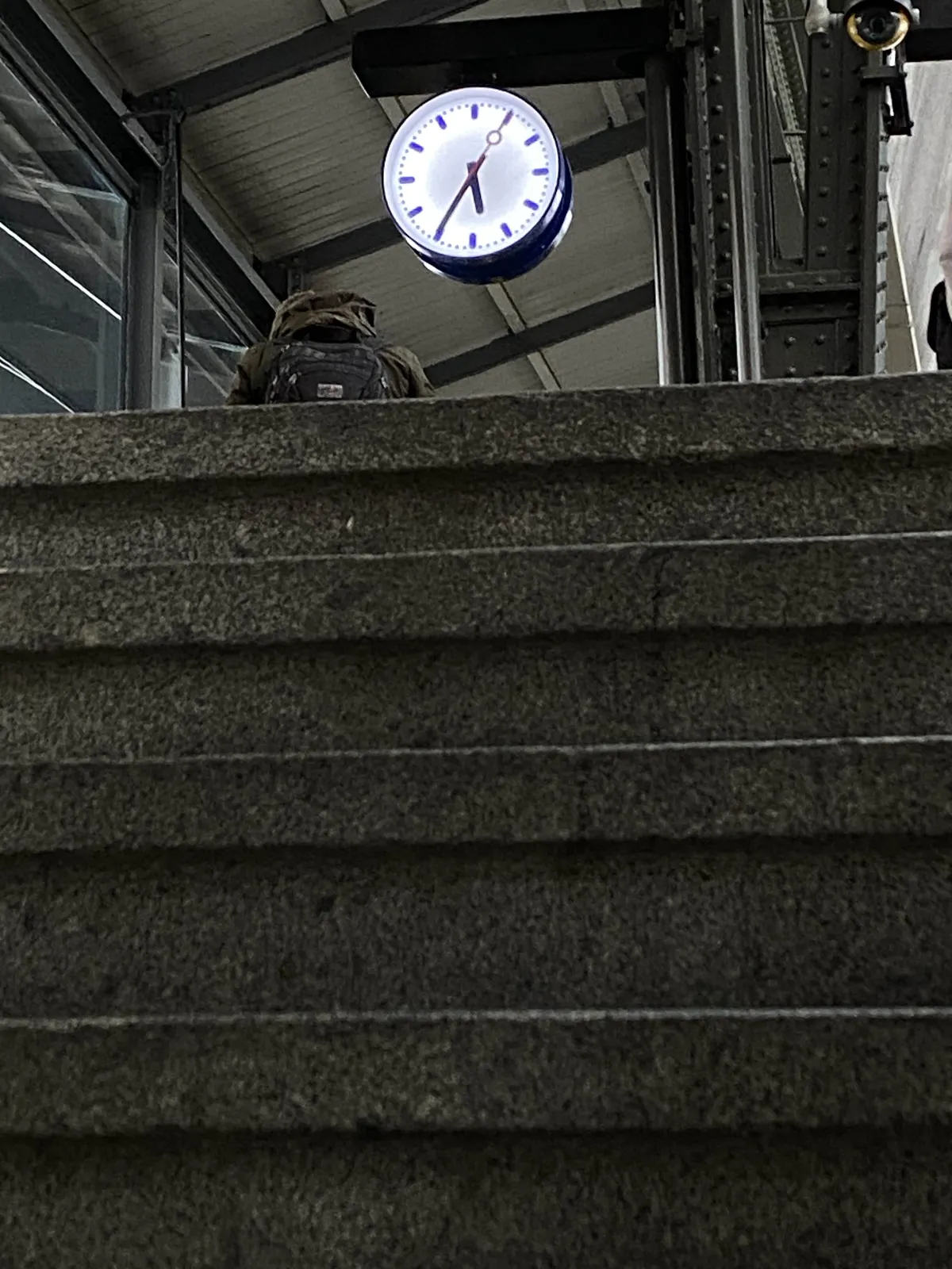 The camera is very close to some concrete stairs. When you look up, over the stairs, there’s a clock. It is 25 minutes to 6.
