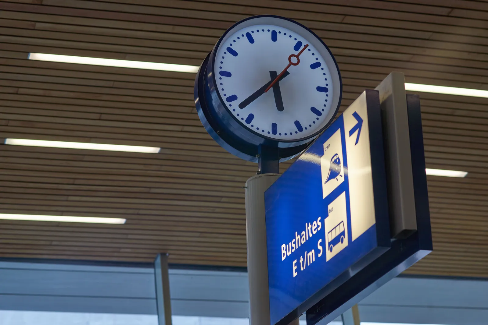 A clock on a pole above a sign that points at the bus stops. On the clock it is 37 past five.