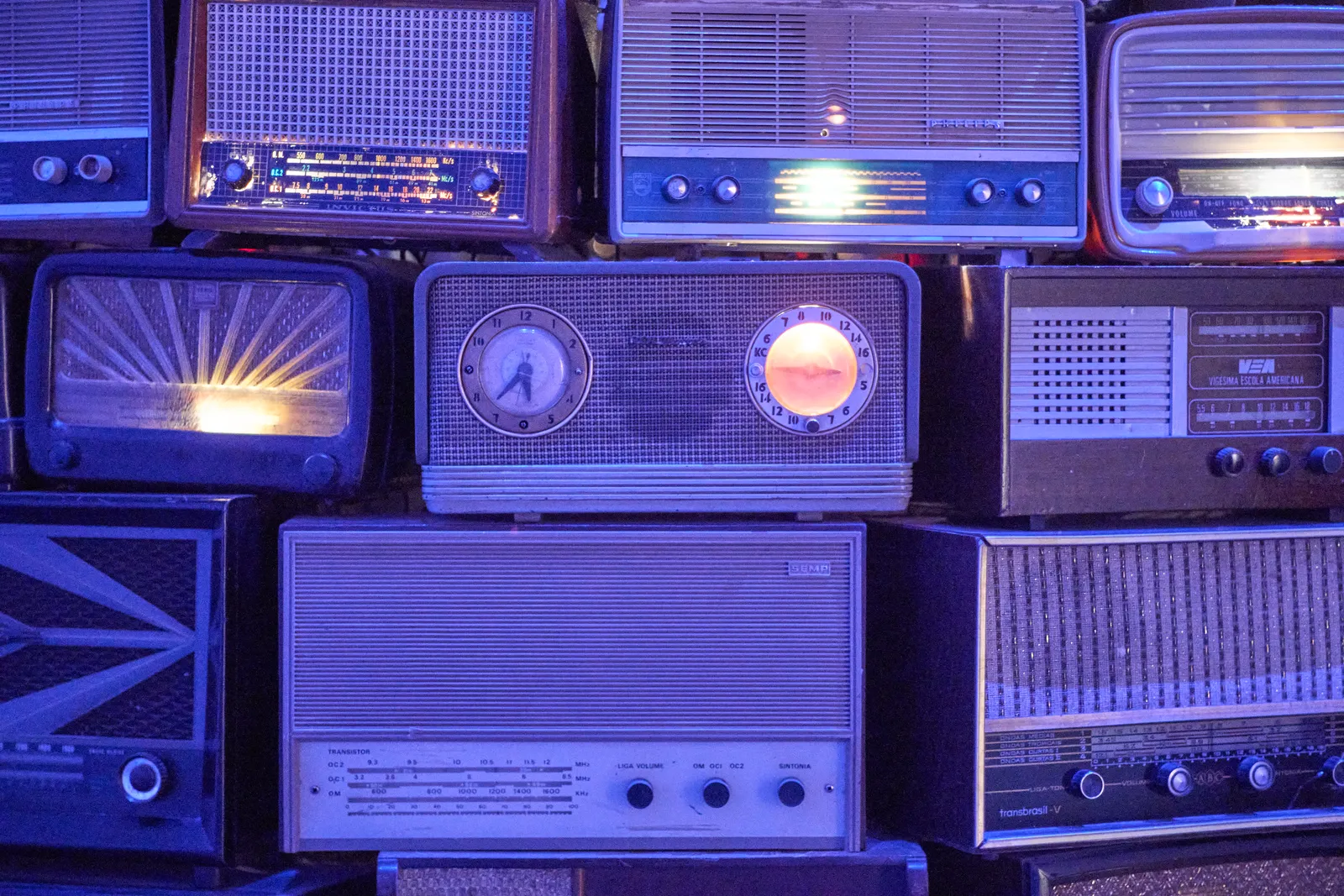 A neat pile of quite a few, old transistor radios. One of these radios also has a clock, which points at 37 minutes past five.