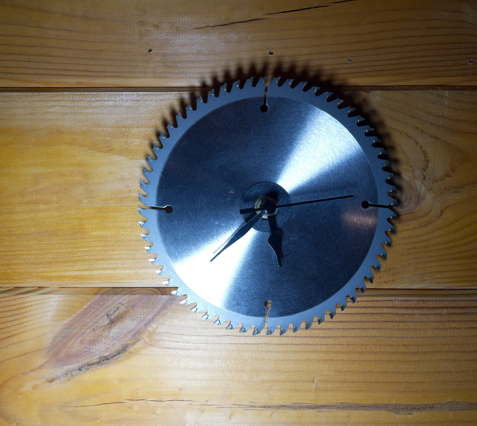 A clock made from a circular saw blade, on a wooden wall. It points at 22 minutes to 6.