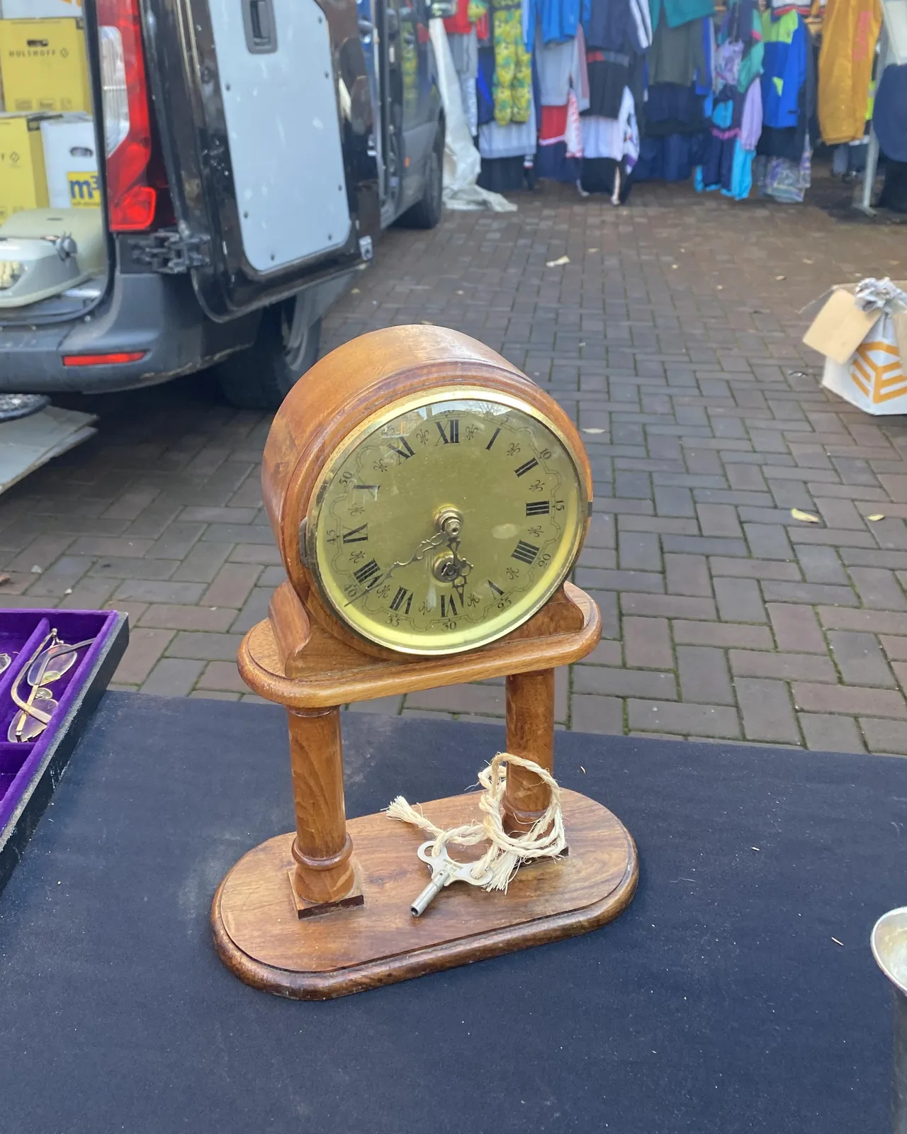 In the background there are second hand clothes for sale, in the foreground there’s an old wooden clock that points at 21 minutes to 6.