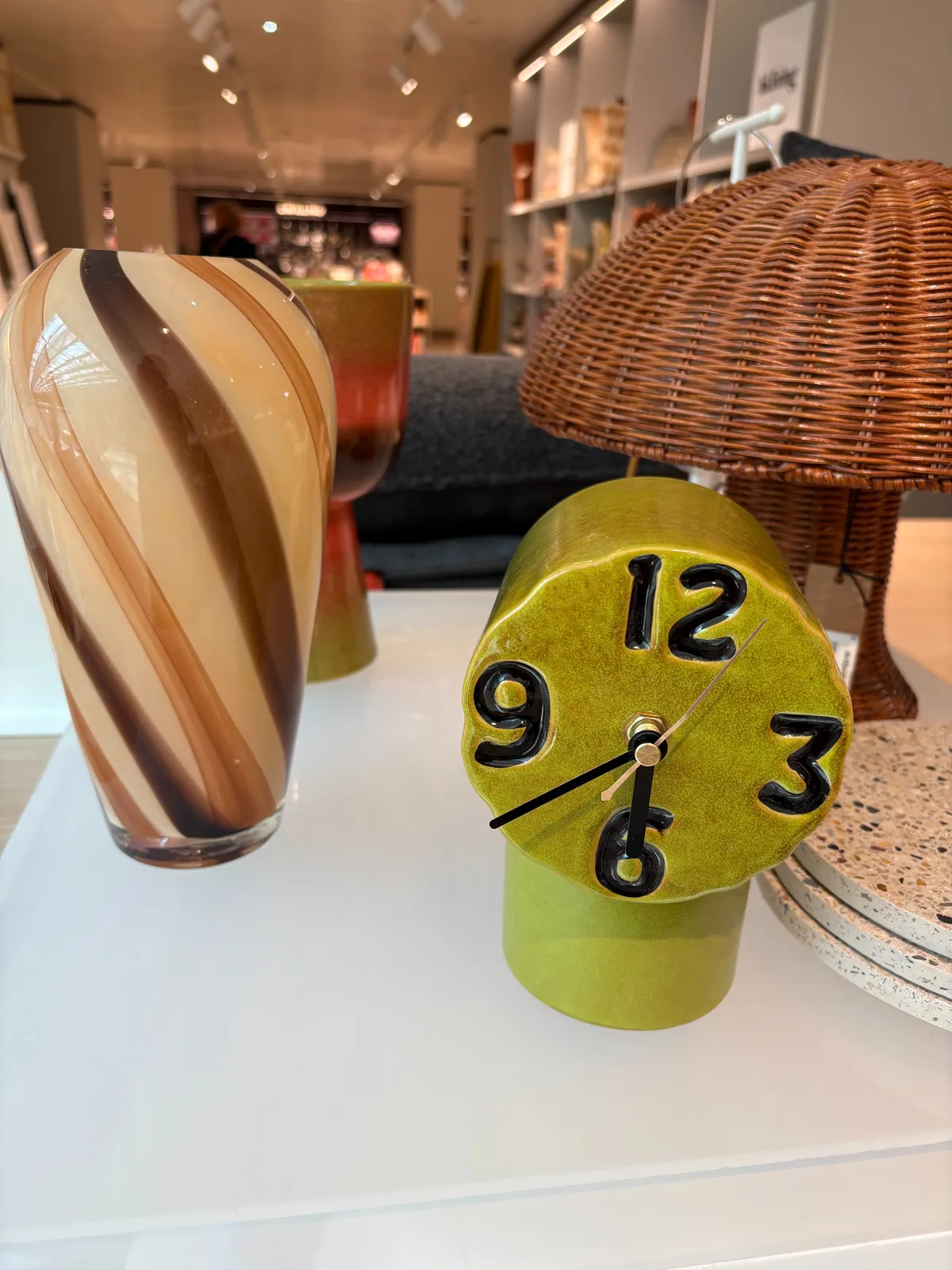 A green ceramic clock on display, next to a colourful glass lamp, and a rattan lamp. It’s unclear what the clock points at. The hour hand points straight down, while the minute hand points at 20 minutes to the hour. So it’s either 20 minutes to 6, or 20 minutes to 7