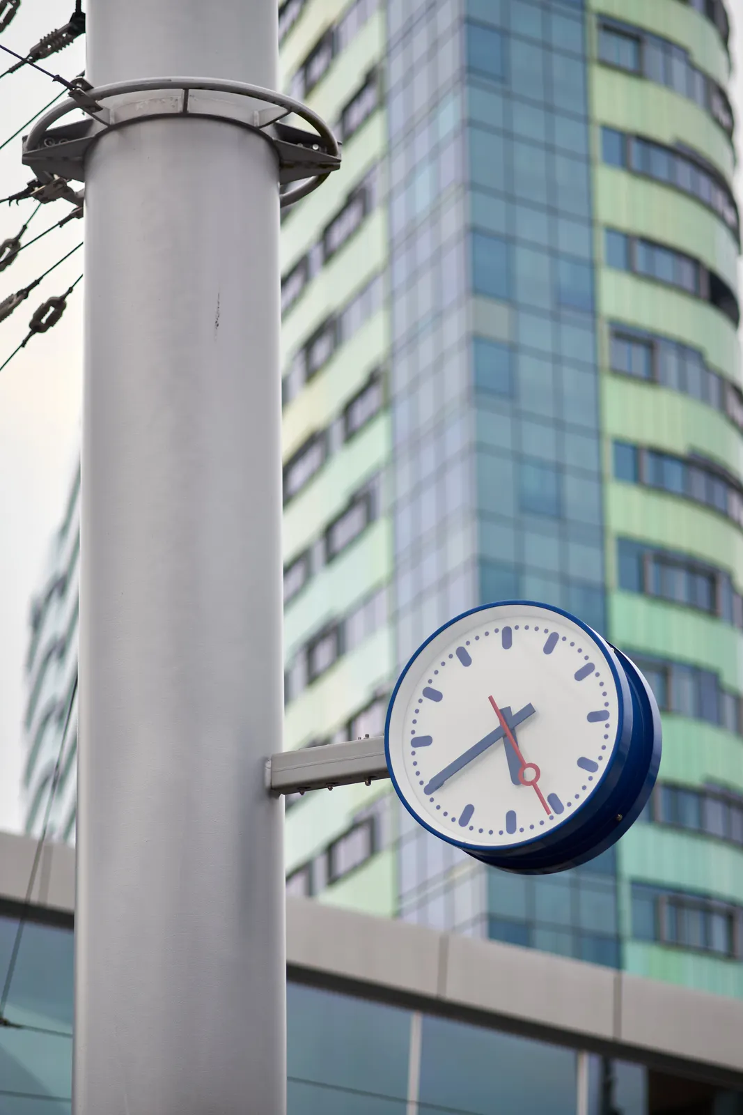 A clock on a pole. In the background there’s a high rise building. On the clock it is 20 to six.