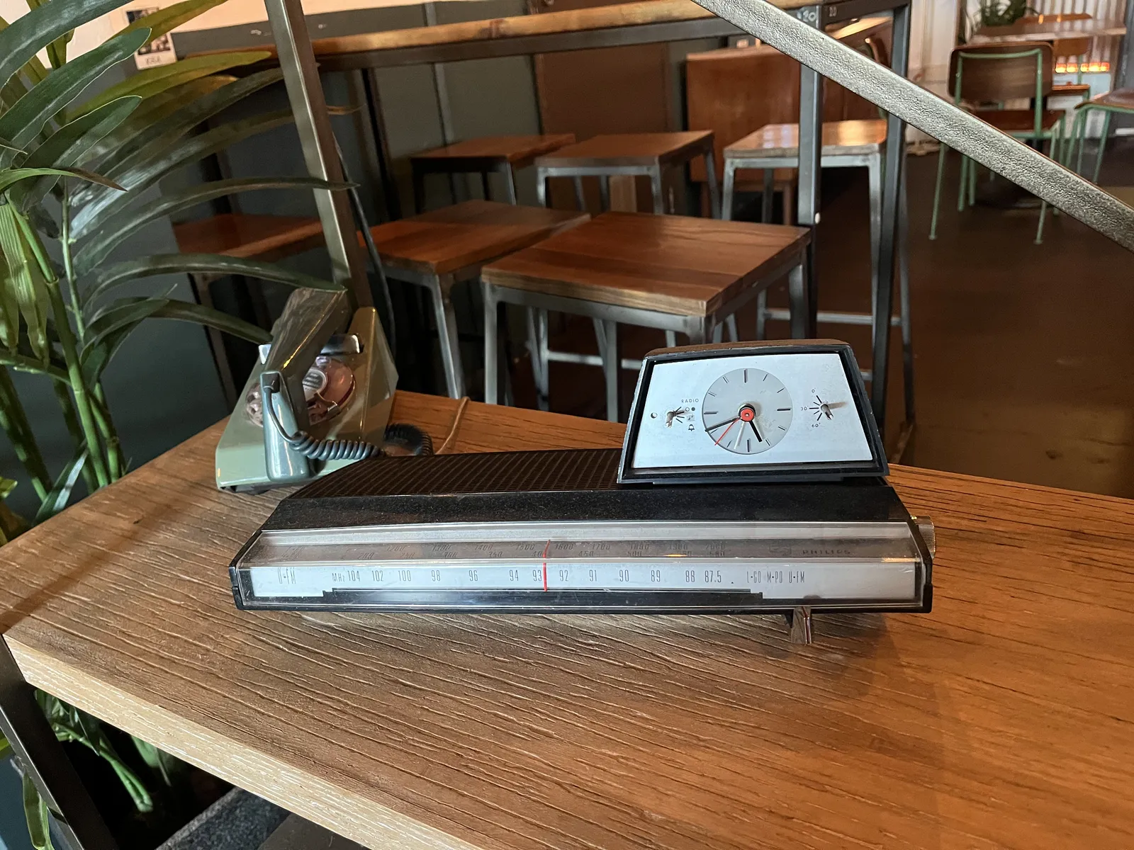 On a wooden shelf, there’s an old, green rotary phone, and some sort of device, it might be a radio, with an analogue clock on it. This clock points at 19 minutes to six.