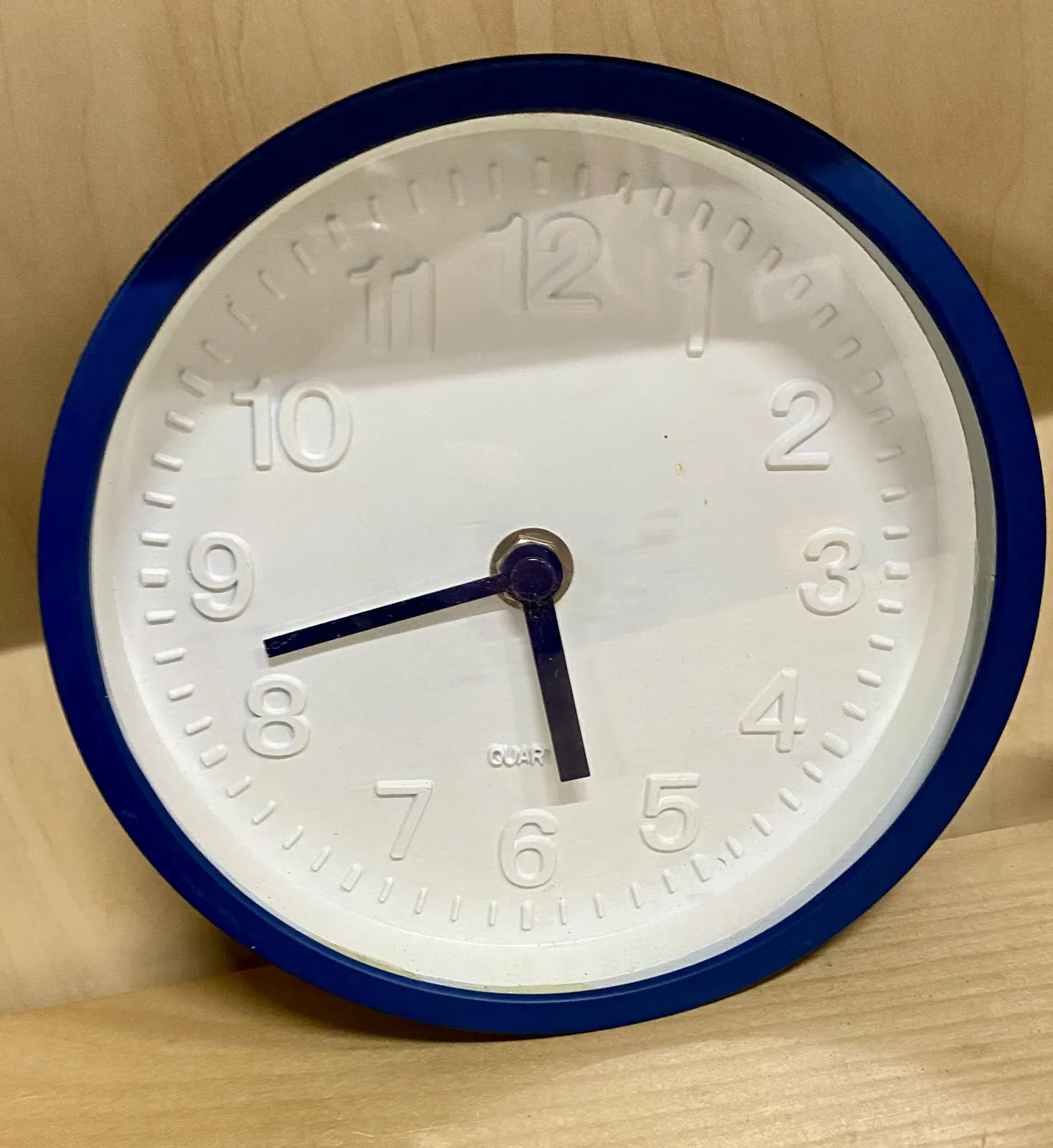 A white round clock with a dark blue border. The numbers are in relief on the face. It points at 18 minutes to 6.