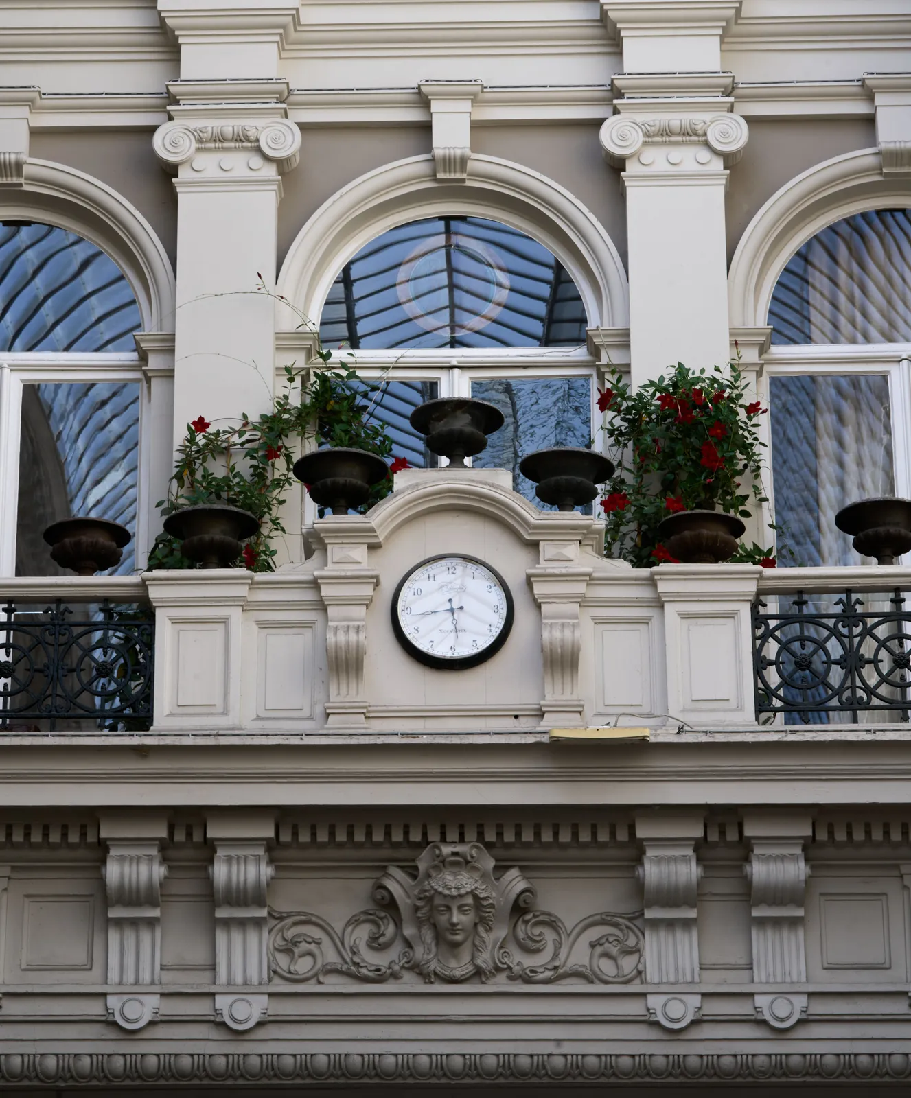 A neo classical balcony with flowers in pots, and a clock. This clock points at 17 minutes to 6.