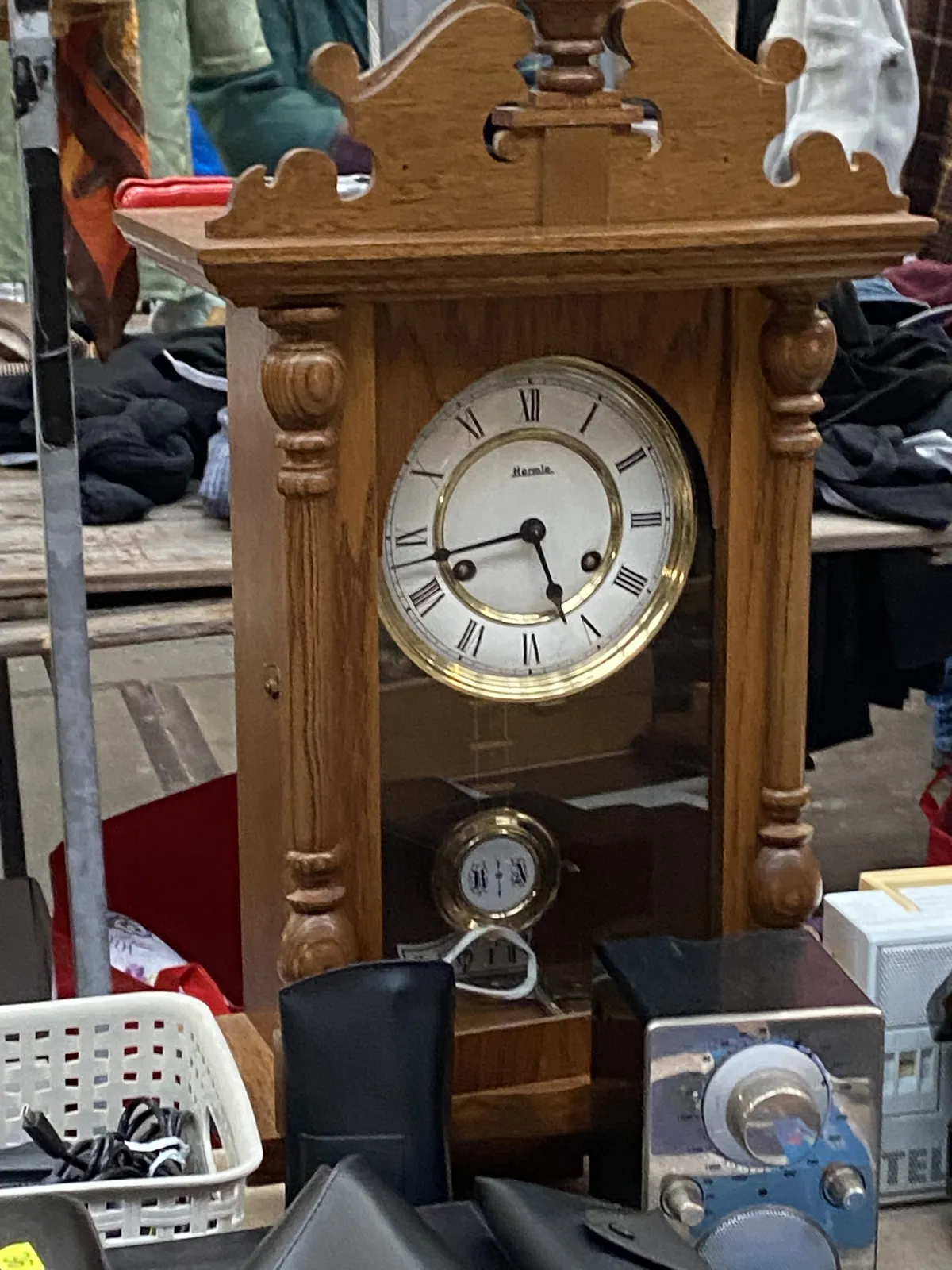 A wooden clock, standing among other stuff on a flea market points at 17 minutes to 6.