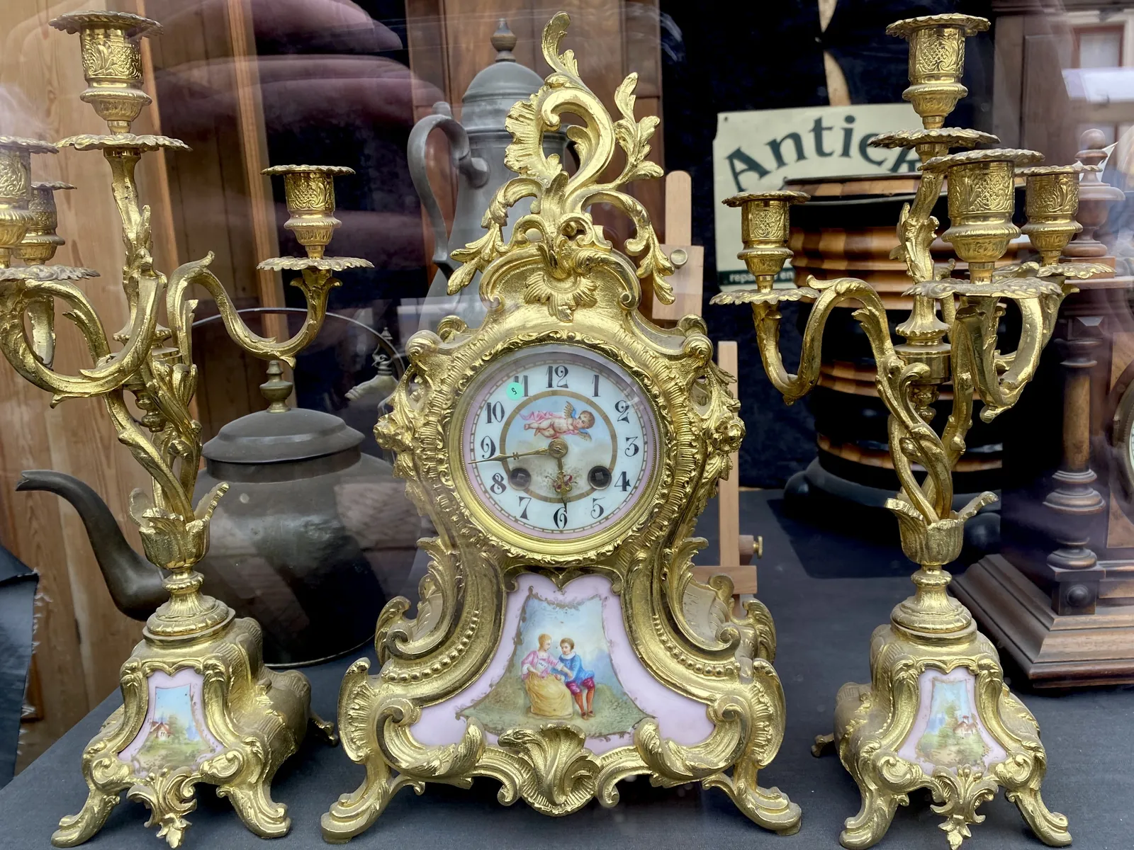 A richly ornamented golden clock, with two accompanying candlestands. A cherub and a couple in love are painted on the clock. It points at 17 minutes to 6.