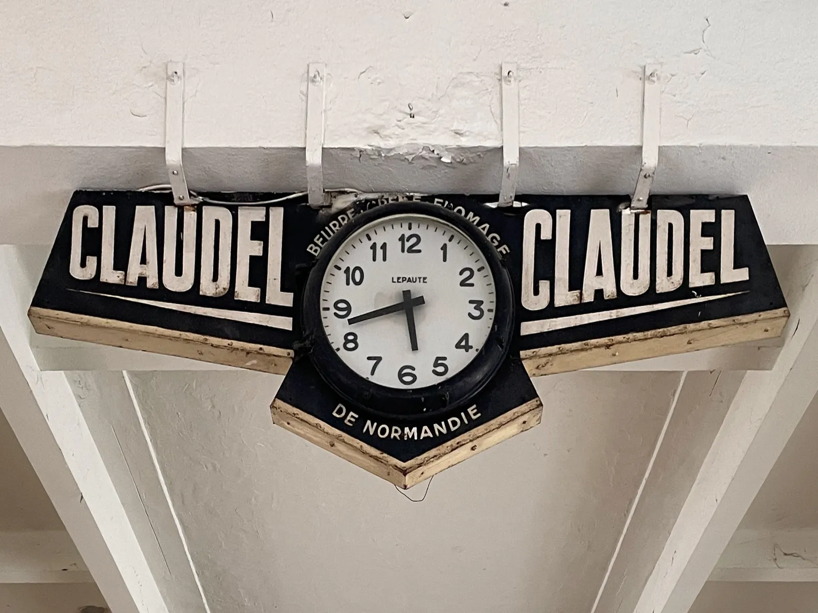 A black clock hanging from a white concrete ceiling. It says Claudel and Claudel on both sides, and Beurre, creme, fromage de Normandie around the clock. The clock is made by Lepaute, and it points at 17 minutes to 6.