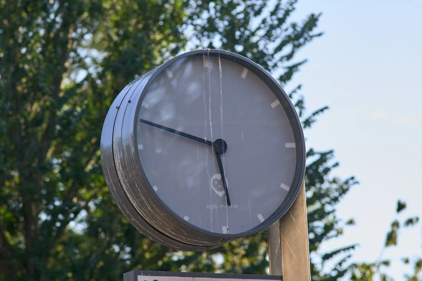 A gray clock against a bright blue background with a blurry tree. There’s a white stripe running down the clock. It points at 12 minutes to 6.