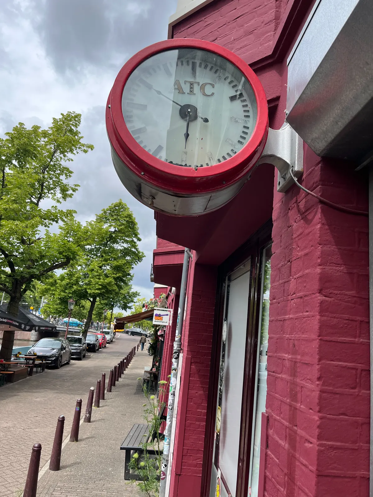 There’s a clock attached to this facade that’s painted red, in a street with trees, some parked cars, next to a canal. The frame of the clock is red as well, and it has the logo of a company called ATC on its face. There are three crosses, or exes, below each other inside the letter T, which may very well indicate that this is an Amsterdam based company. This makes sense, when you look at the canal. The clock points at 10 minutes to six.