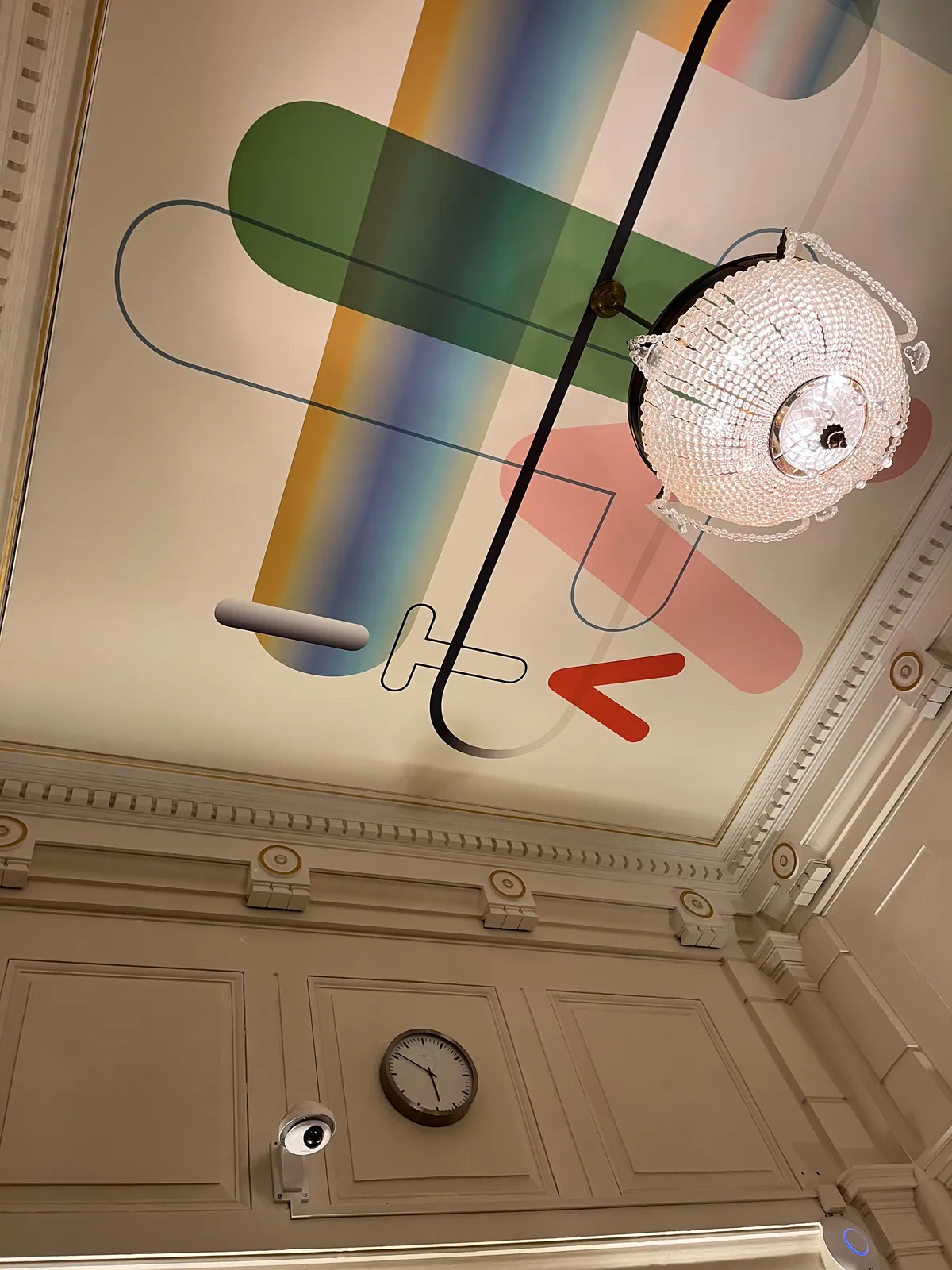 A very colourful, illustrated ceiling (a ceiling indeed). There’s also a chandelier which looks a bit like an eye. There’s another eye-like object, but that’s a spy camera. Above this camera hangs a clock which points at 9 minutes to 6.