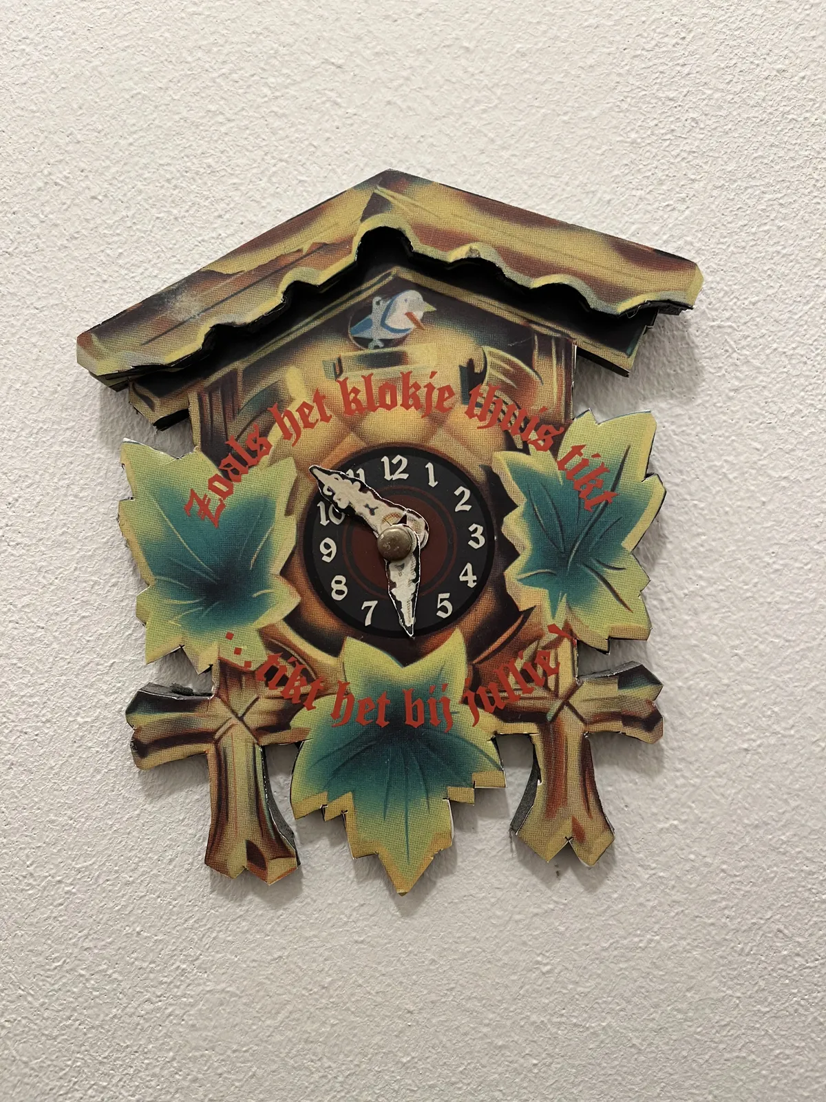 A cut out print of a cuckoo’s clock, with the phrase on it, in Dutch: zoals het klokje thuis tiks tikt het nergens, which translates to: The the clock ticks at home, it ticks nowhere. The paper hands of this clock point at 8 minutes to six.