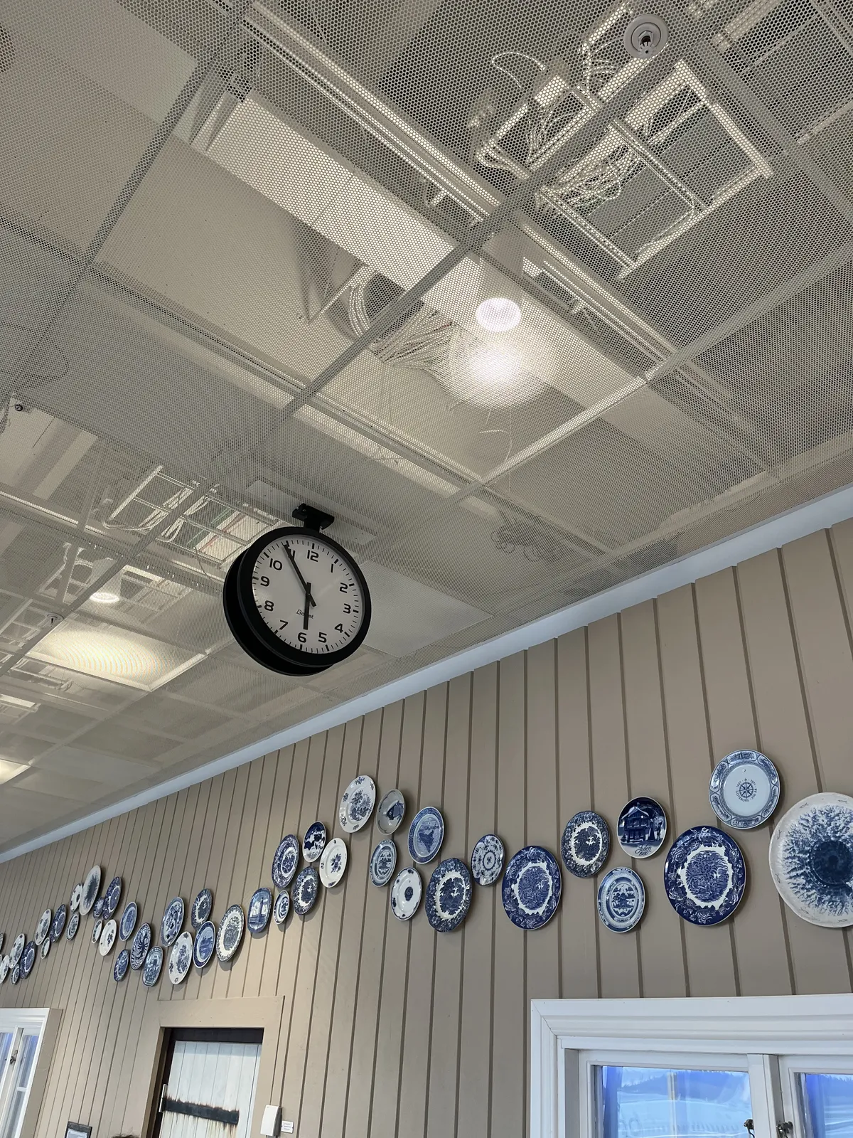 Many, many blue and white plates, all with different illustrations, form a wave on a beige wall. The ceiling is made from metal plates with holes in them. You can see the electrical wiring behind them. A clock is hanging from this ceiling. It points at 6 minutes to six.