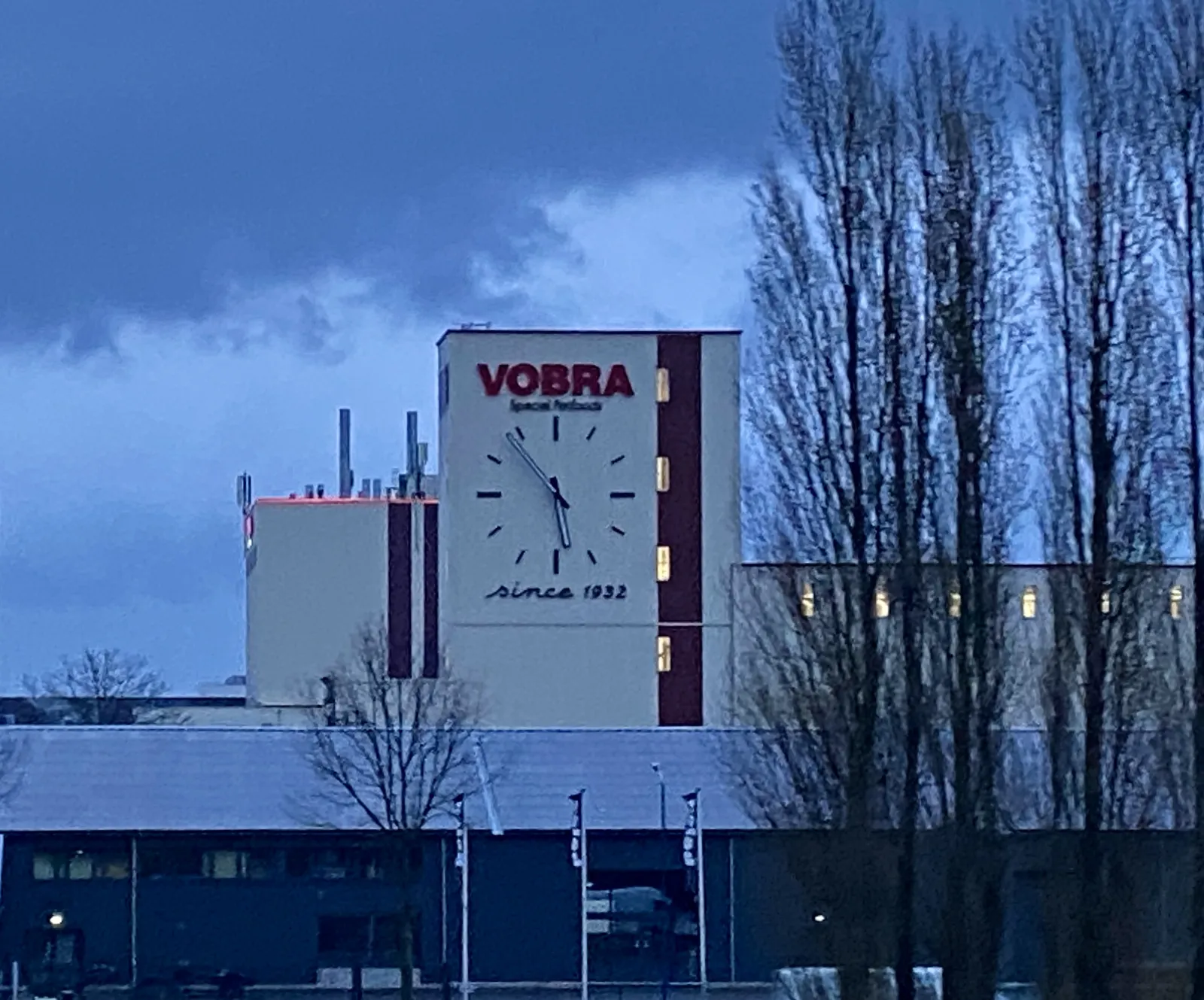 A very large clock on a factory building. It says Vobra, special something (unreadable), since 1932. It points at 6 minutes to 6.