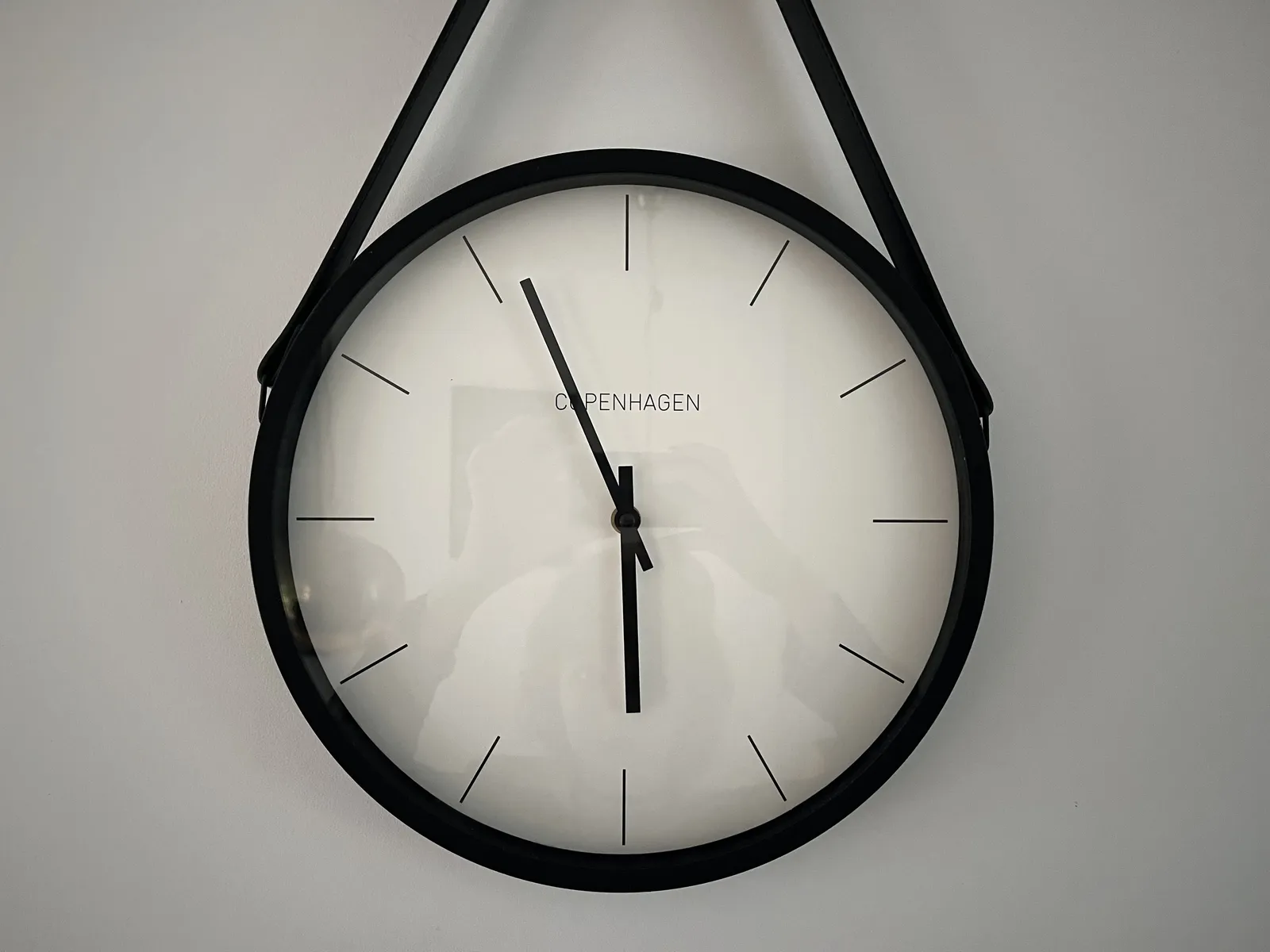 A modern, plain, black and white clock. The word Copenhagen is printed on its face in uppercase in a plain, thin sans serif type face. There are no numbers on it, just lines indicating the hours. It points at approximately 4 minutes to 6. The long hand is hiding the letter O, so to be honest, it’s not entirely clear if it realy says Copenhagen. In the reflection in the glass that covers the clock, the photographer can be seen taking the picture by holding their camera above their head.