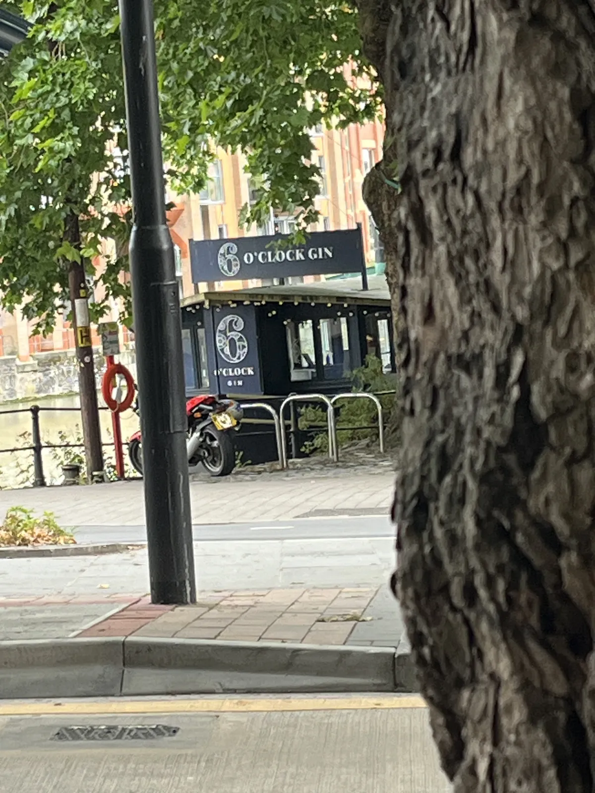 Peeking past a tree, past a lamp post, to a bridge over a canal or a river. Next to this canal is a sign, it could be on a boat, that reads 6 o’clock gin.