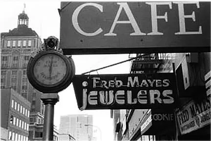 A street clock, next to a cafe, and next to a sign that says Fred Mayes Jewelers, points at one past six.