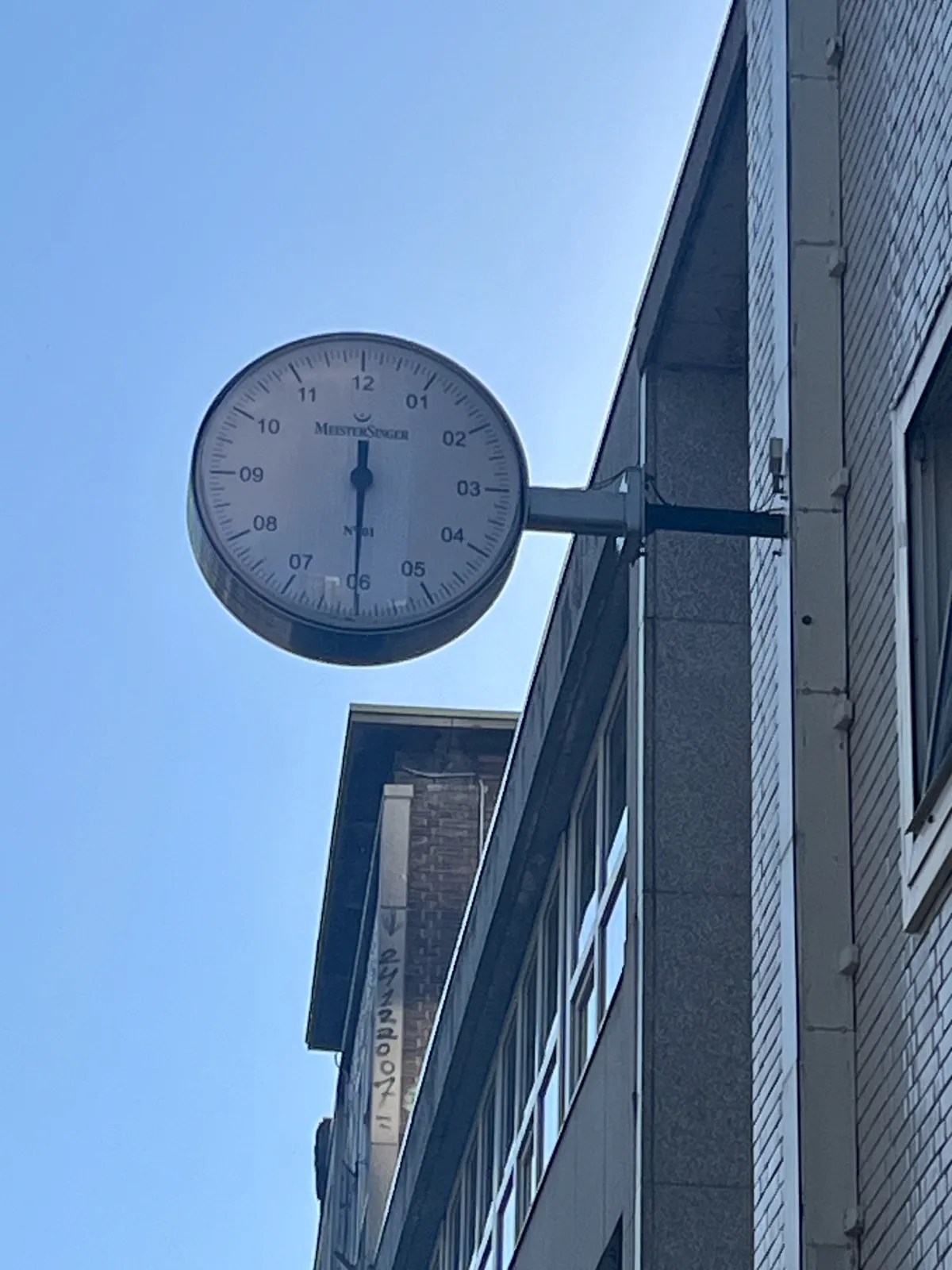 A clock attached high on the facade of a building. It has just a single hand, which points down, just slightly past its southermost point. Which means that it is approximately 2 minutes past six.