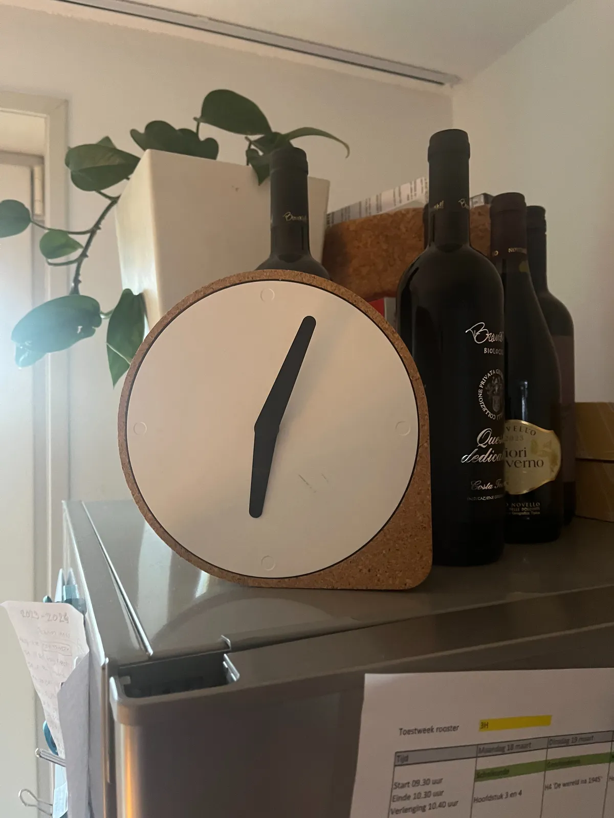 A plant, a few bottles of red wine and a clock standing on a fridge. The clock points at approximately 3 minutes past 6.