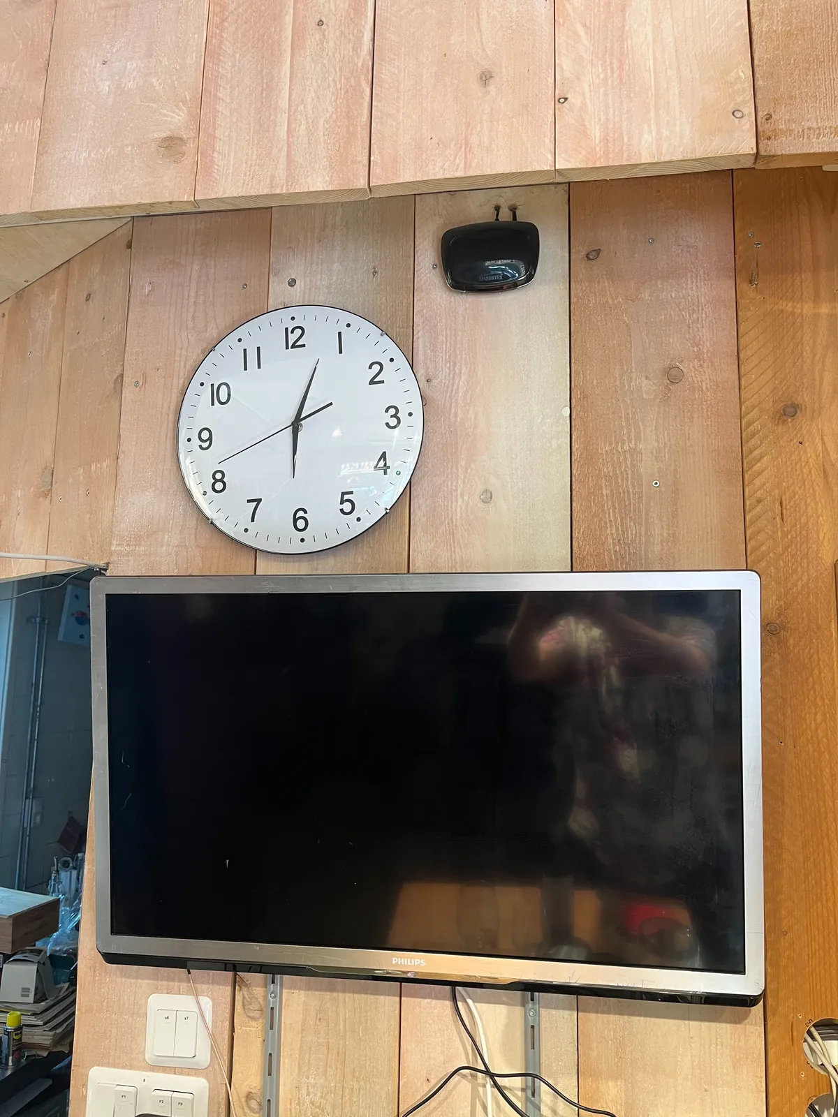 A simple, white, modern wall clock, hanging on a wooden wall, above a black TV screen. It points at 3 minutes past six.