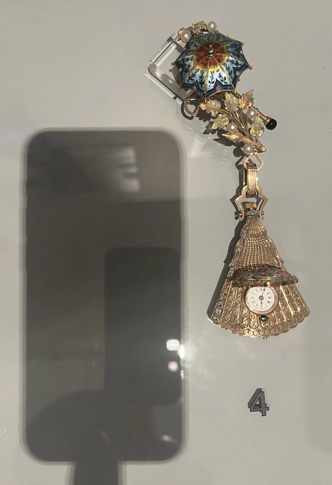 In a glass showcase there’s a richly decorated, golden, brooch, with a strikingly colourful umbrella on it. It’s decorated with pearls and other precious stones and it has a clock attached to it, which is placed on a golden, half open folding fan. The picture is taken with a phone, which is placed on the glass top, and the photographer is touching the phone with one finger. The shadow of this phone and finger take up the complete left side of the picture. The brooch with the clock is on the right. The clock points at 4 minutes past six.