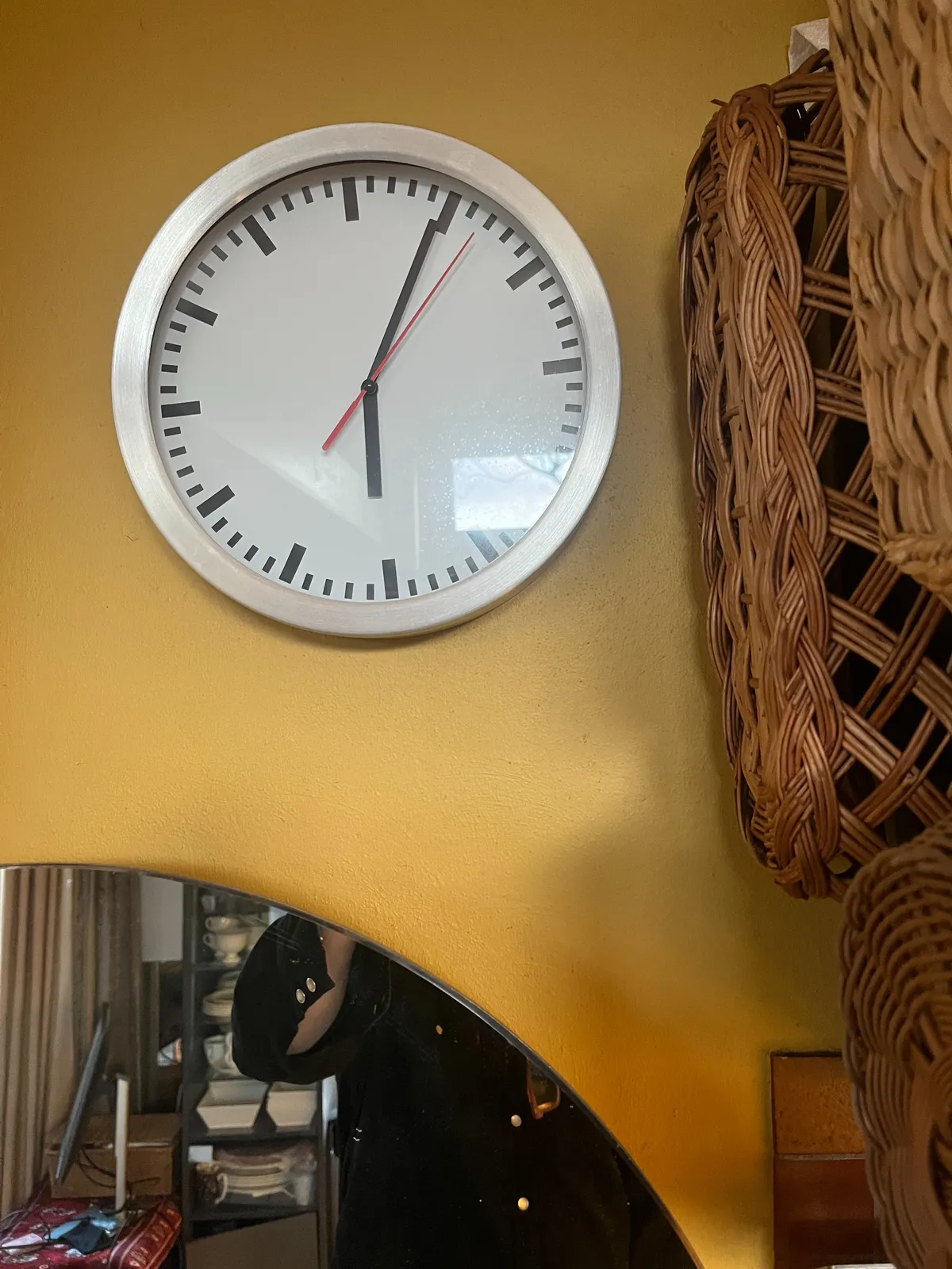A simple wall clock on a yellow wall. There are bread baskets and a mirror around it. The clock points at 4 minutes past 6.