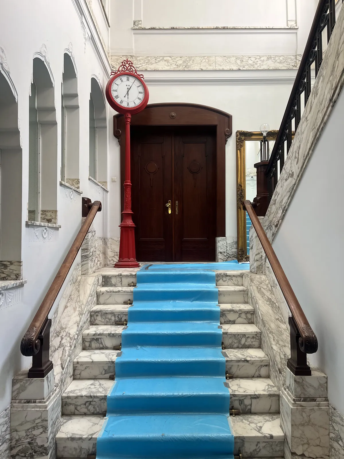A blue carpet on luxurious, old, marble stairs. In the corner there’s a red pole with a red clock on it which points at five minutes past six.
