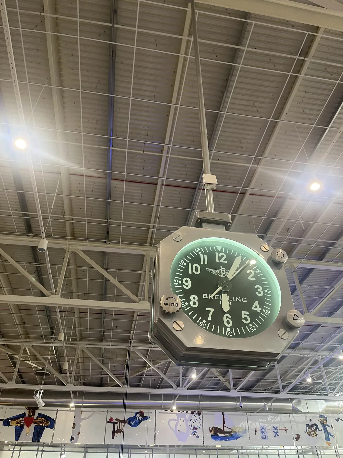 A clock is hanging from the ceiling in a very high, moder hall. It looks a bit like a wrist watch, it says Breitling on its face, and it points at 5 past six.