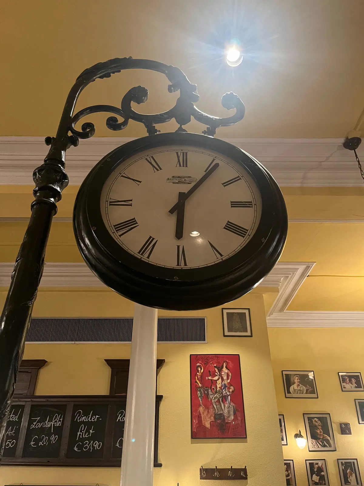 A large clock hanging from a pole inside a restaurant. It points at 6 minutes past 6.