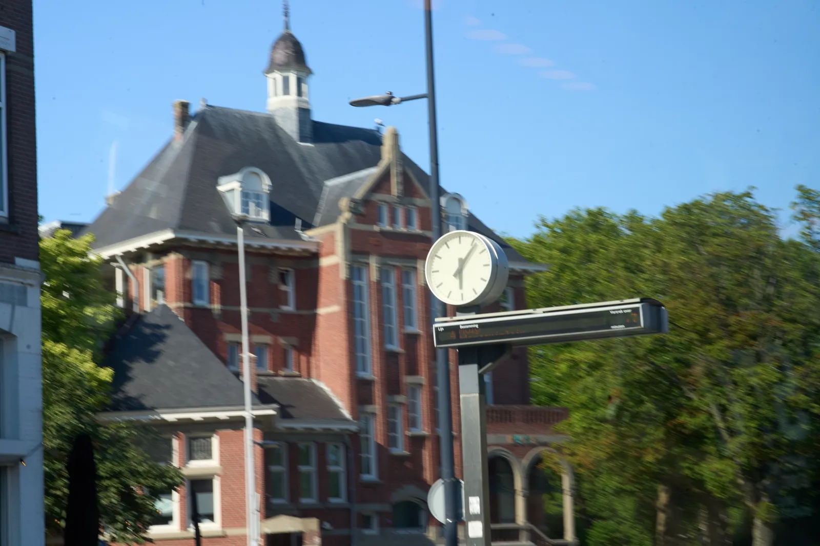 A clock on a pole, with a thin, wide digital display below it. The clock is in focus, the background, with an old building and some trees, are blurry. The clock points at 6 minutes past six.
