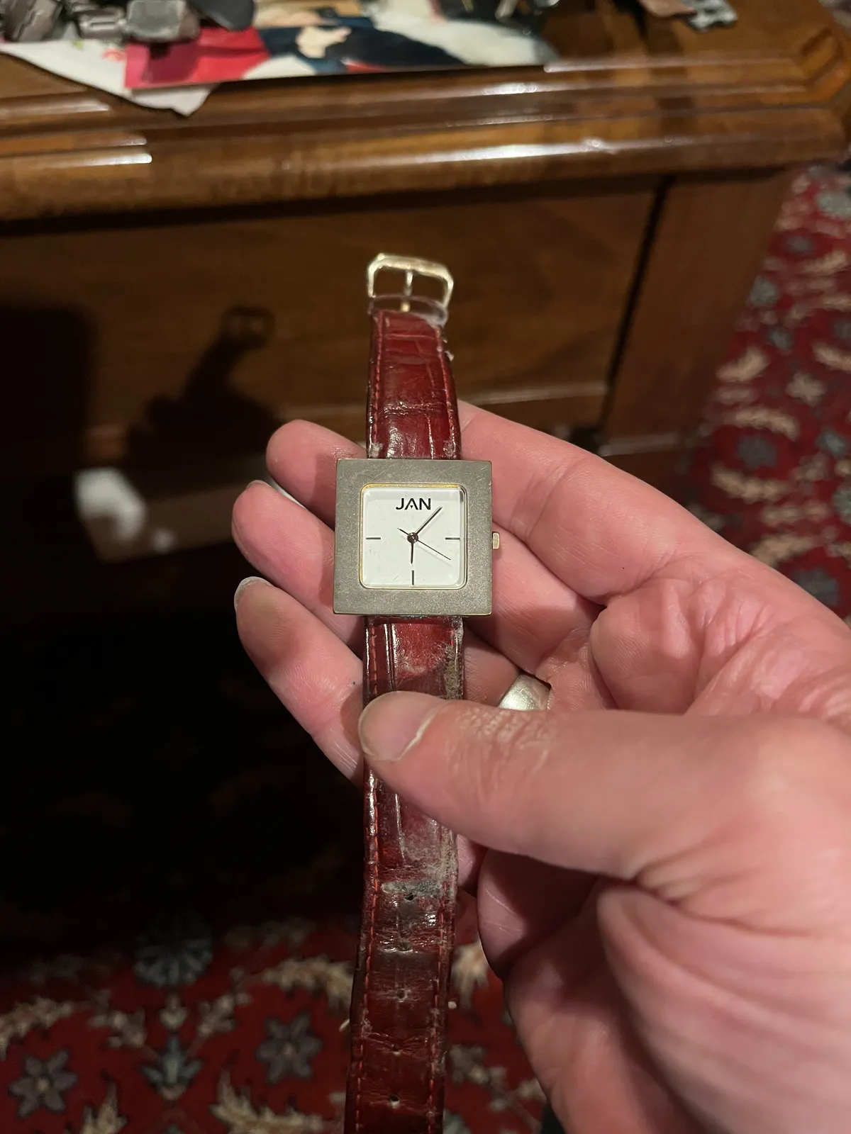 Someone is holding an old watch in their hand. In the background, blurry, an old Persian rug. The watch points at 14 minutes past one. It says JAN in its face.