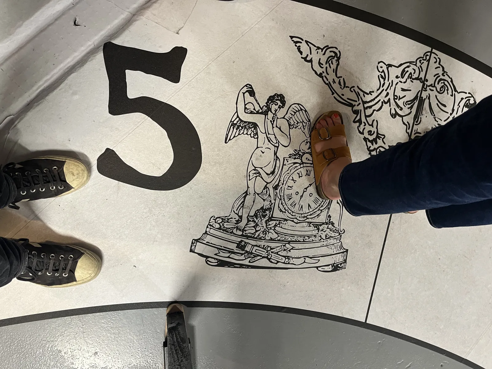 People standing on a painted floor. Their feet can be seen. One of the things painted on the floor is an image of an older, decorated mantel clock, which points at seven minutes past six.