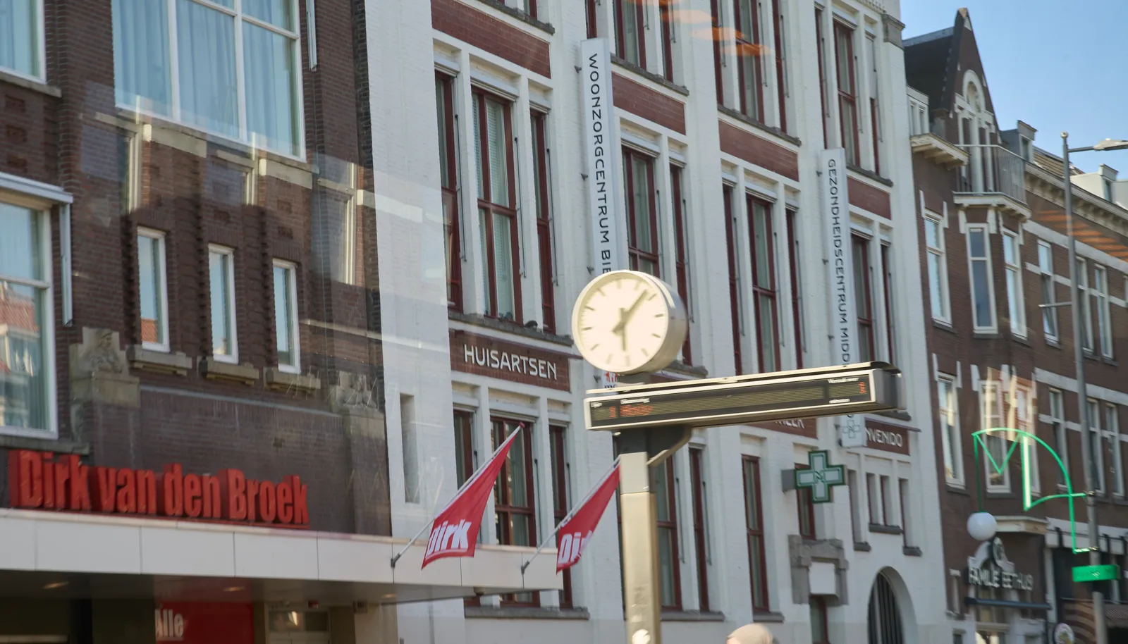 A clock on a pole, with a thin, wide digital display below it. The clock is out of focus, the background, which consists of a large facade of a Dutch city building, with a health center and a super market in it, is in focus. The clock points at 7 minutes past six.