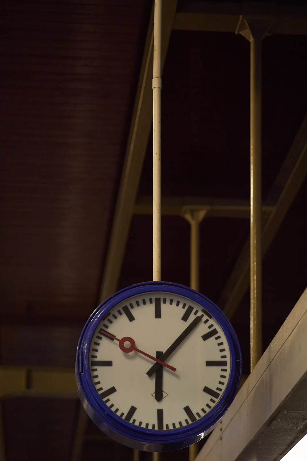 A clock hanging from a pole from a wooden ceiling. The clock is all the way at the bottom of the picture. It points at 7 minutes past 6.