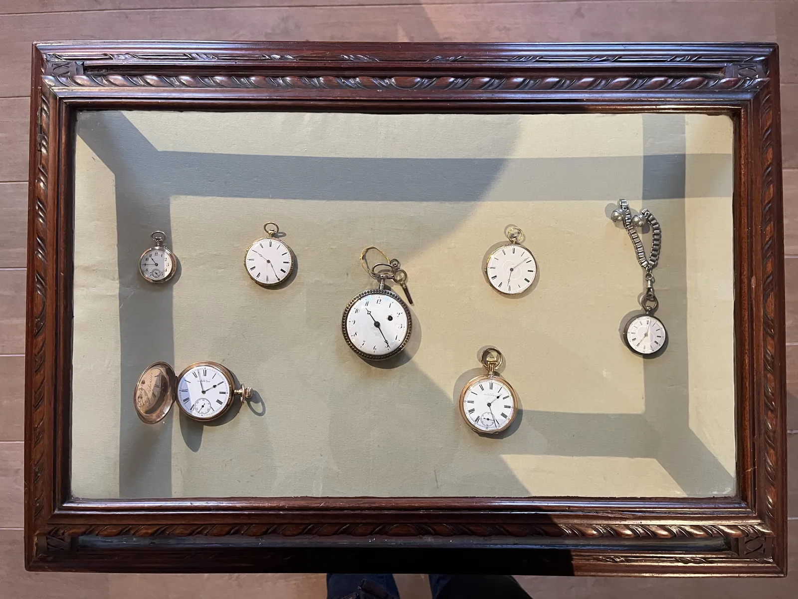 Seven old, delicate looking, silver pocket watches in a display cage. One of the watches opints at 26 minutes past one. Another points at 3 minutes to two. There’s one that points at 8 minutes after six. One of the watches points at 2 minutes past 7 o’clock. There’s one that says that it is 24 minutes past 10, and another that says it is 25 minutes past ten. And finally there’s one small watch that points at a quarter to eleven.  